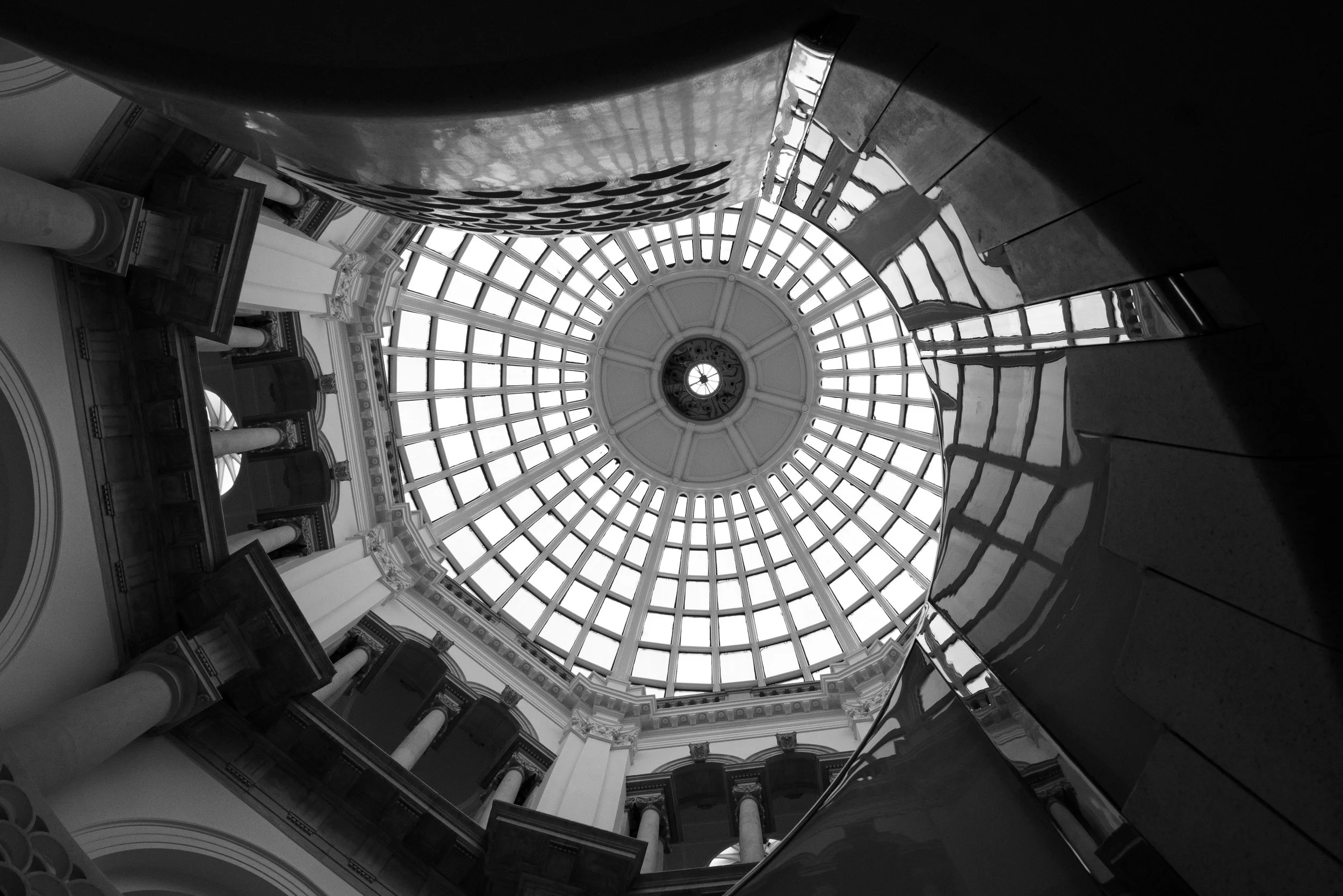 Black-and-white upward view of an ornate domed interior with a radial glass skylight, classical columns, and reflective sculptures.