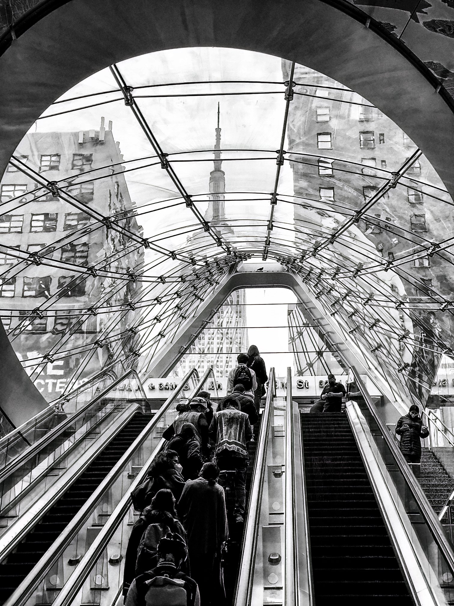 People riding escalators beneath a glass canopy in a busy New York City subway station.