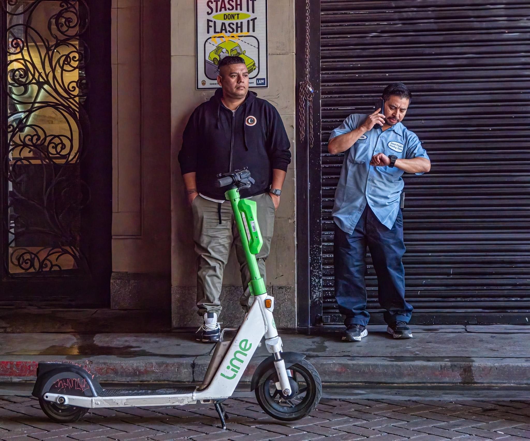Two men stand beside a Lime electric scooter outside a storefront, with one talking on a phone and a vehicle security sign behind them.