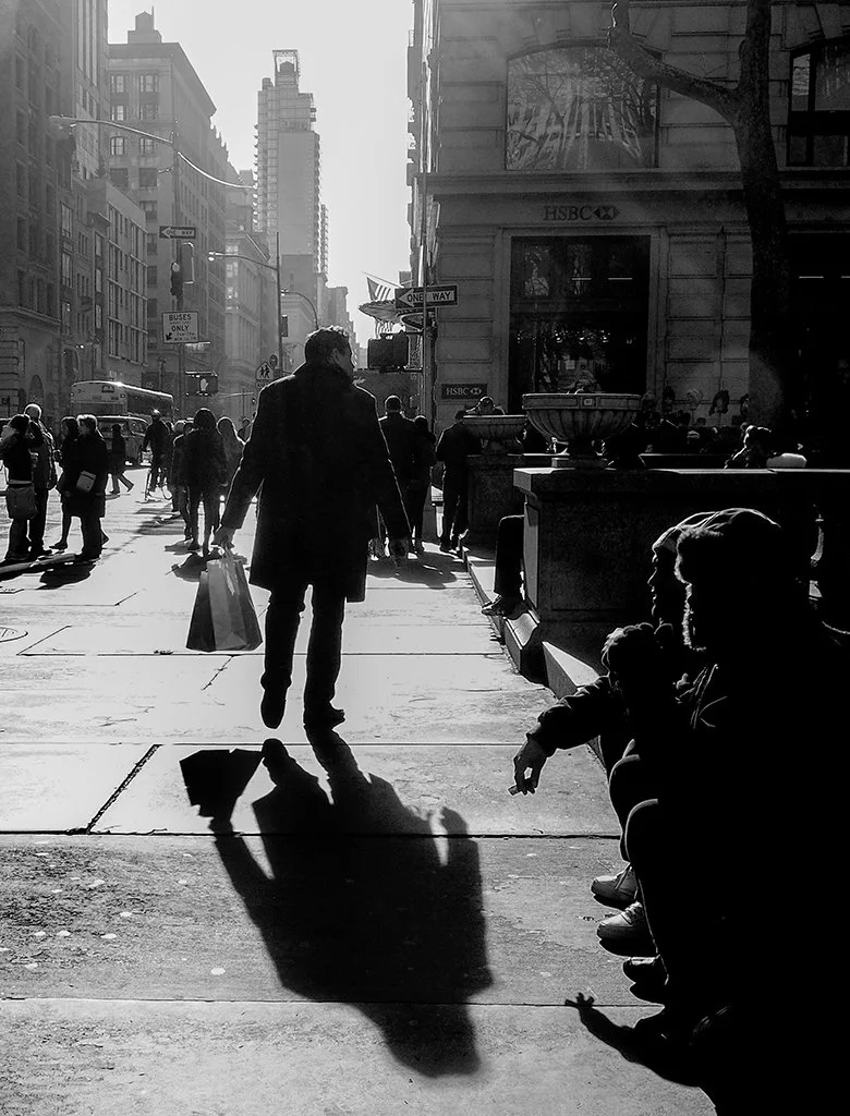 Black-and-white street scene with pedestrians, a city bus, HSBC bank, and long afternoon shadows on a busy urban sidewalk