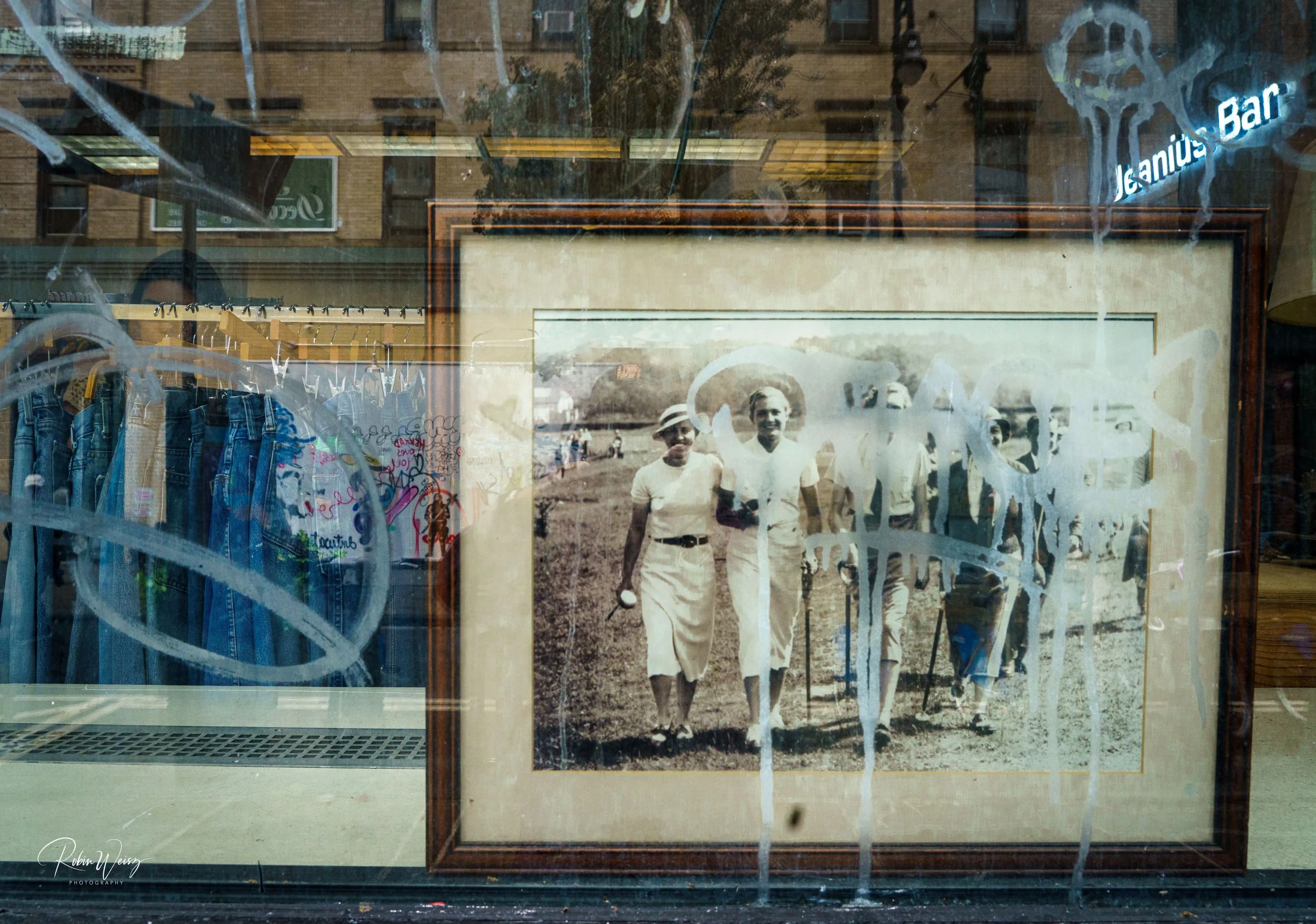 Shop window displaying a framed vintage black-and-white photograph of people in white clothing, with hanging denim garments and reflected street signs.