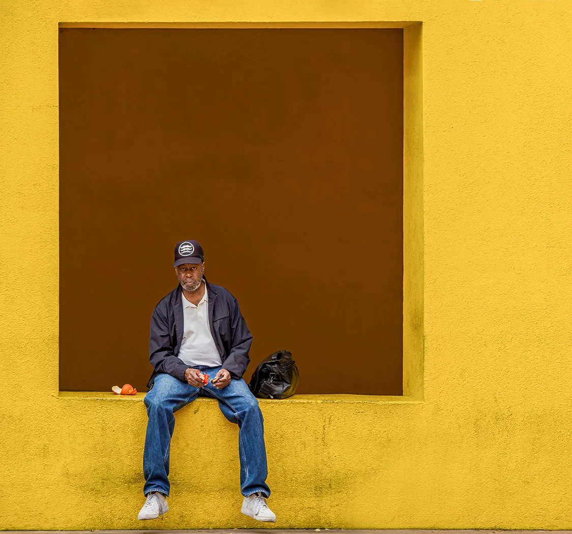 Man in a cap and jacket sitting in a recessed window of a bright yellow wall with a backpack beside him