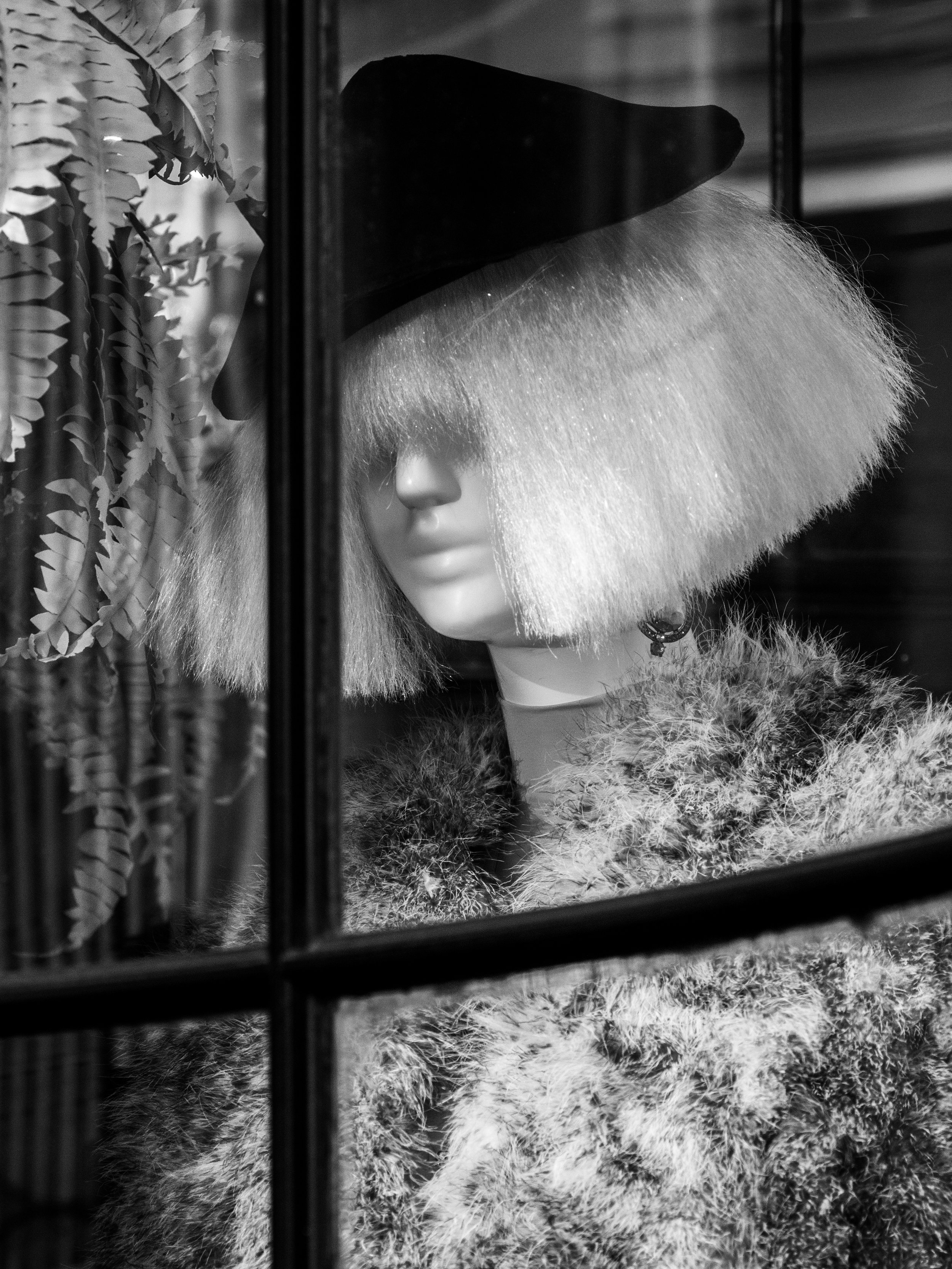 Black-and-white close-up of a woman in a wide-brimmed hat and furry coat behind a window with leafy curtains.