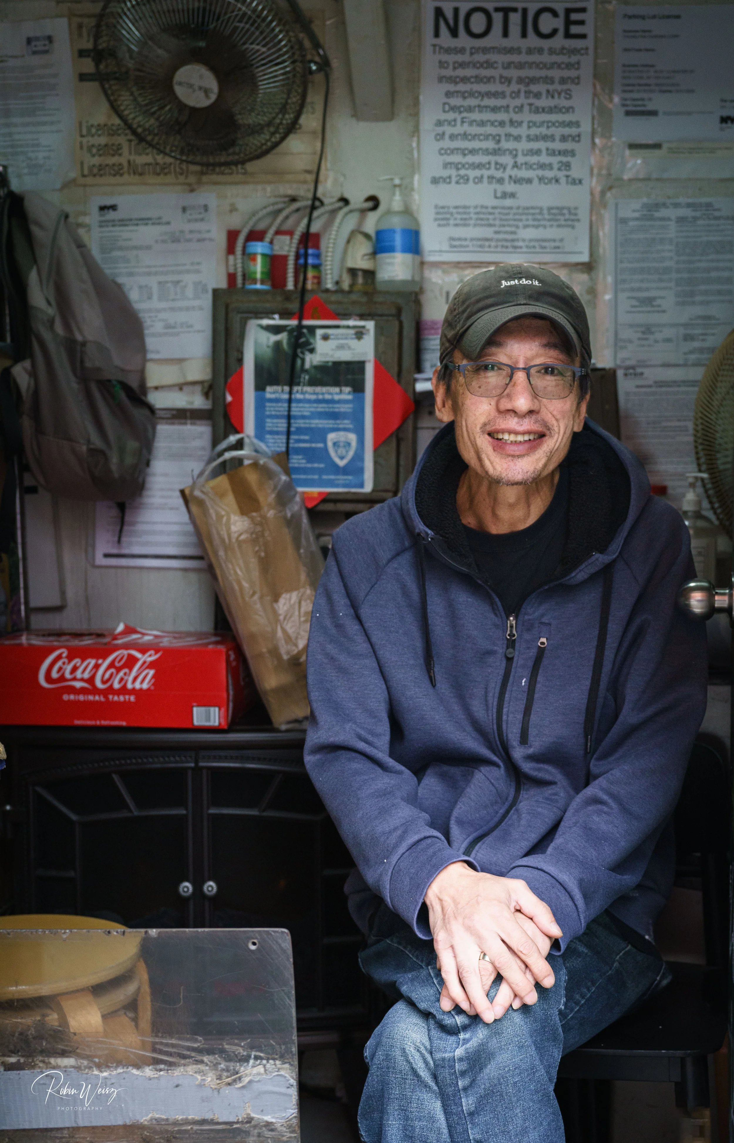 Smiling man wearing glasses, a cap, and blue jacket seated inside a cluttered shop with posted notices, supplies, and a fan.