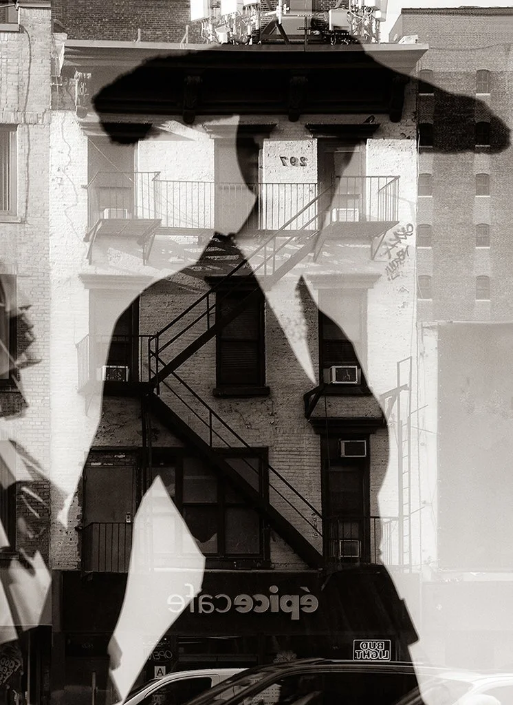 Black-and-white double exposure of a silhouetted person overlaid on a brick urban building with fire escape stairs and storefronts