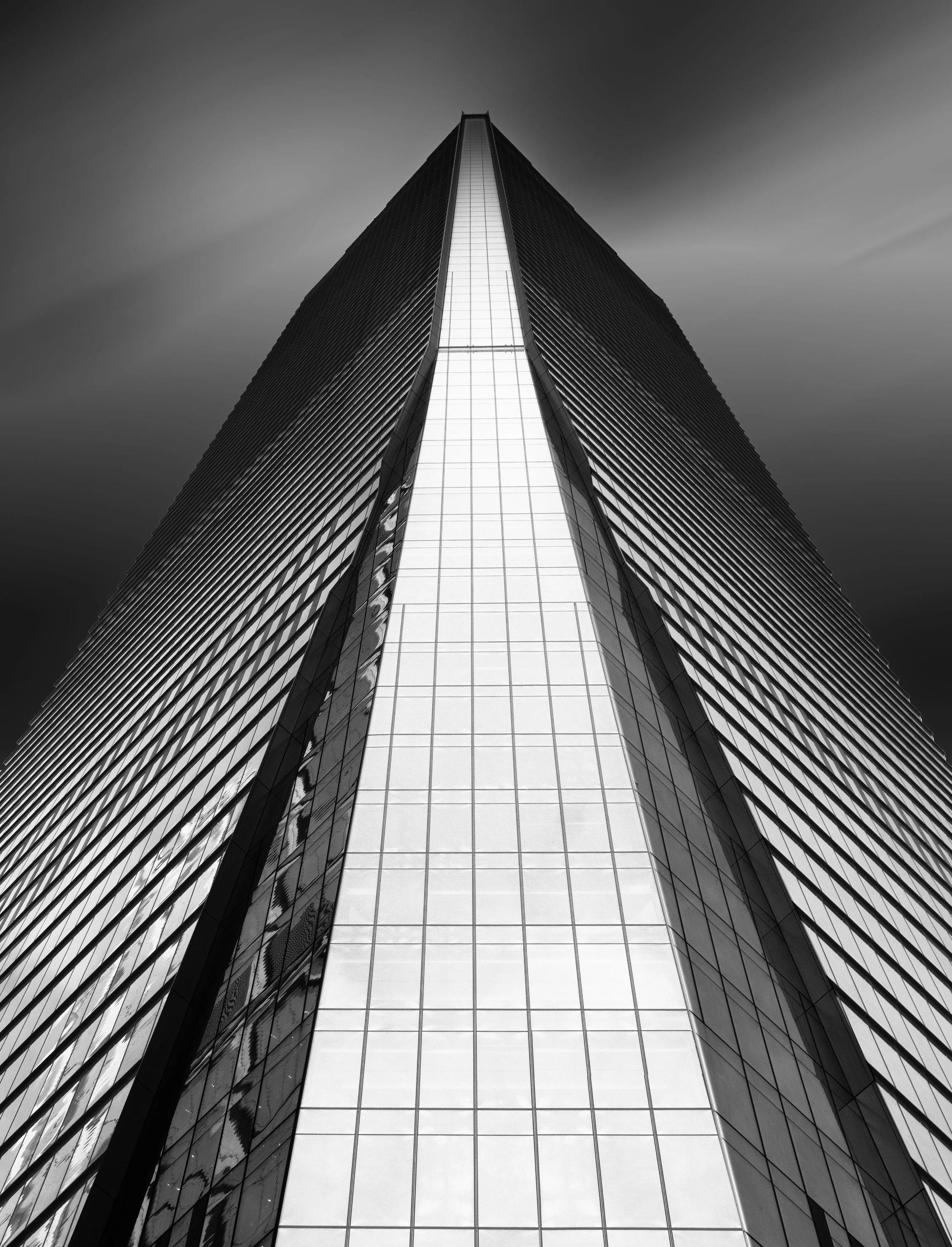 Black-and-white low-angle view of a towering glass skyscraper against a cloudy sky