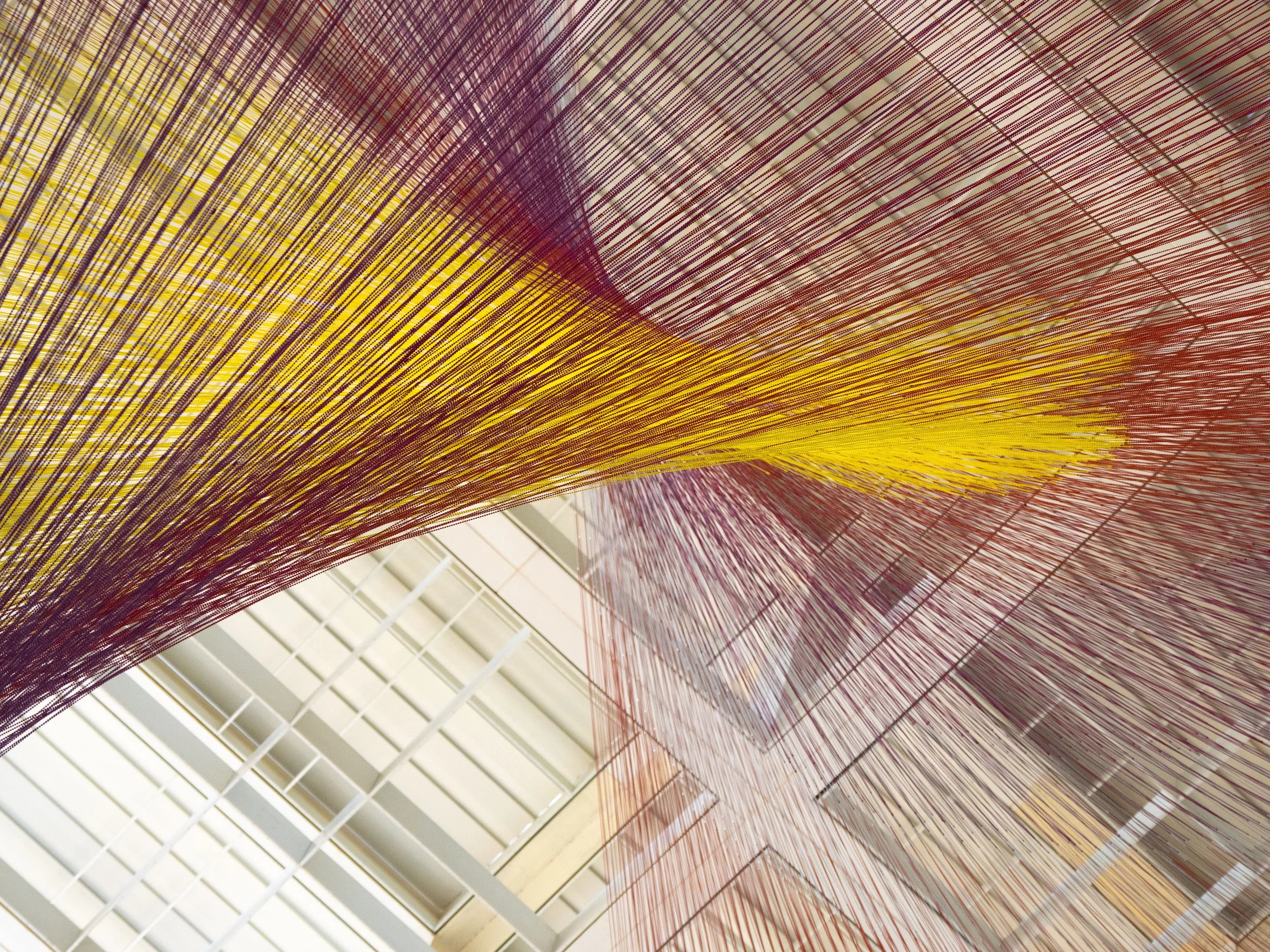 Abstract installation of thousands of purple, yellow, and red suspended threads forming geometric patterns beneath a skylight