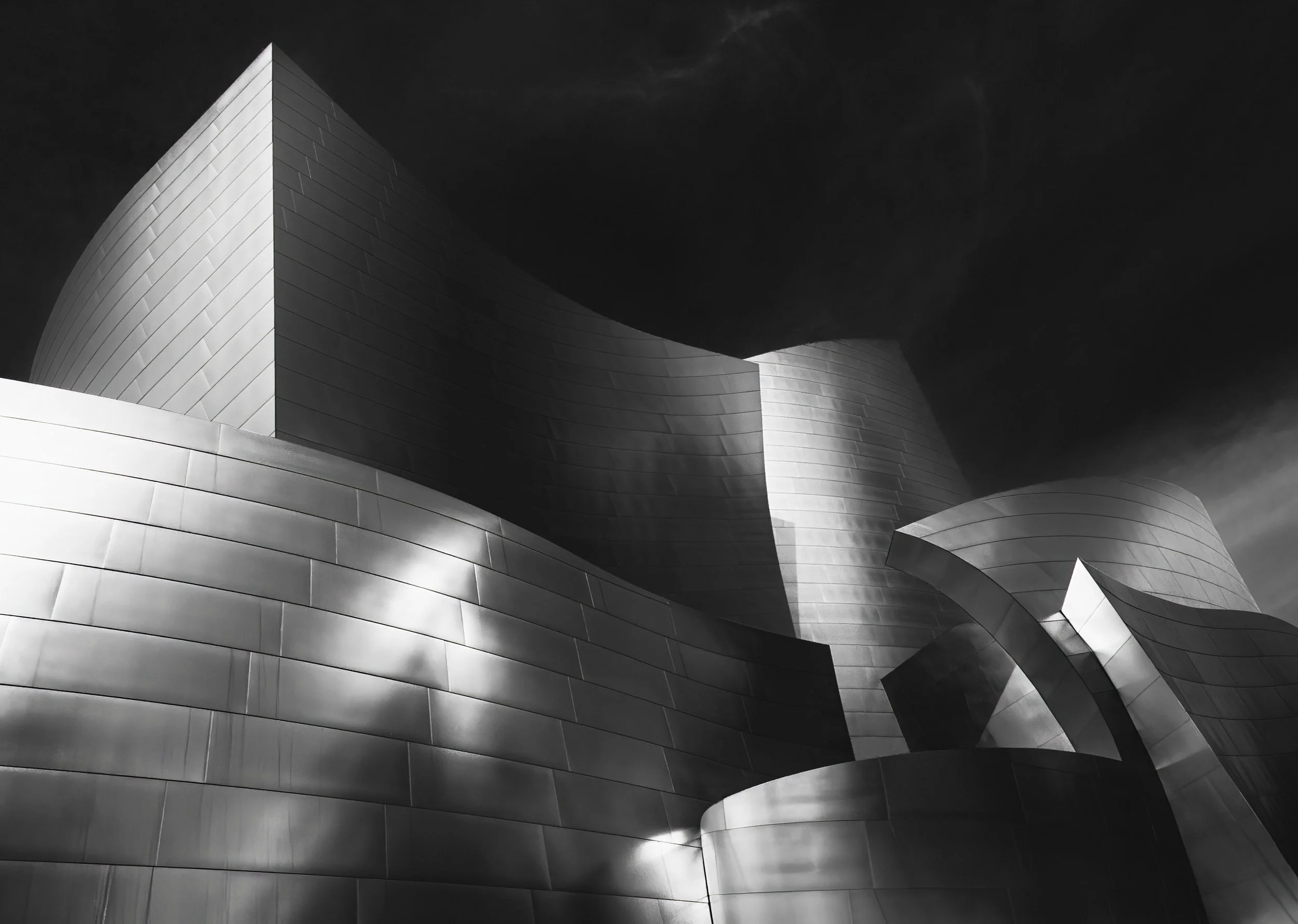 Black-and-white low-angle view of the Walt Disney Concert Hall’s sweeping stainless steel curves beneath a dramatic cloudy sky