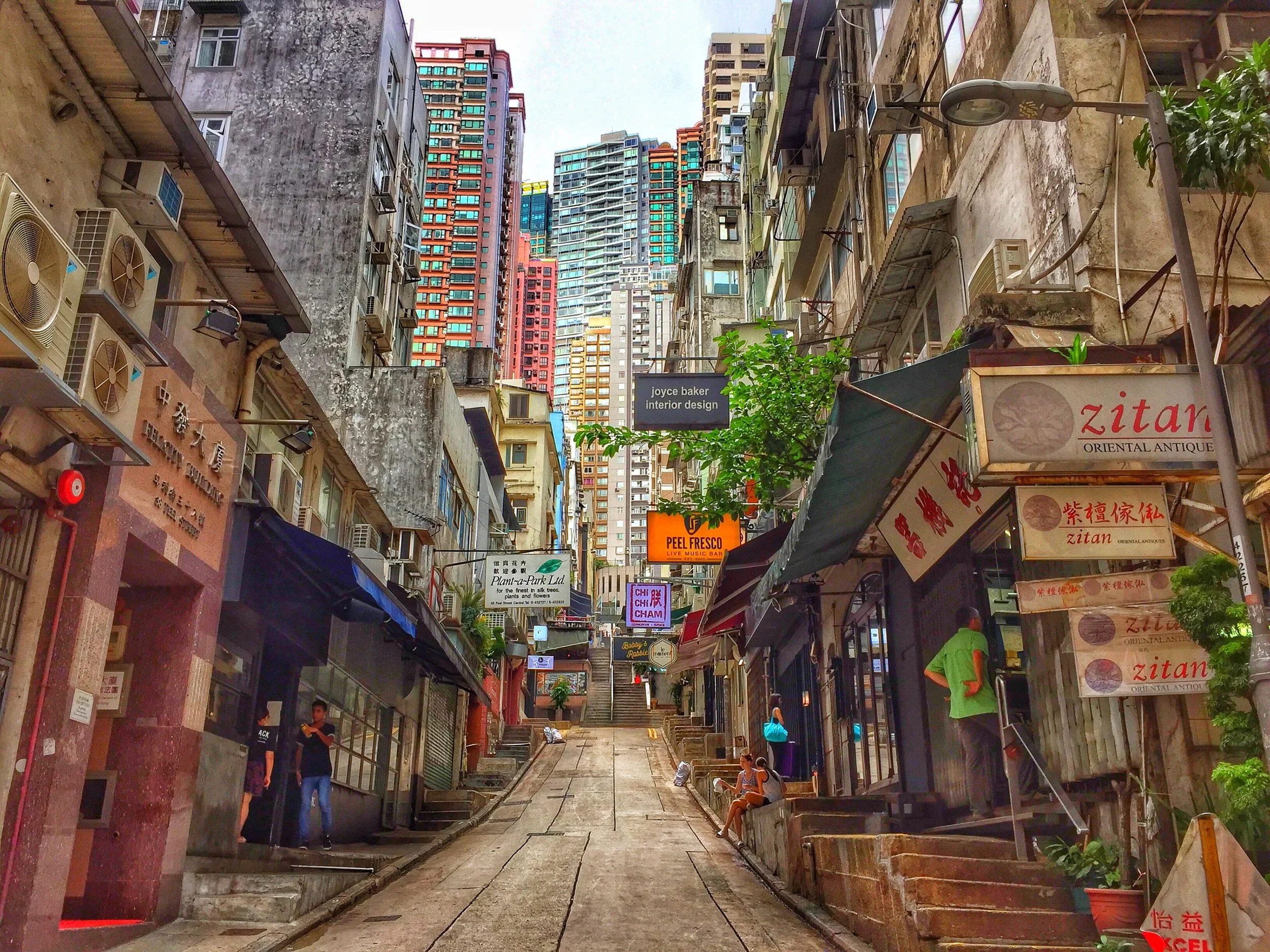 Steep urban alley in Hong Kong lined with shops, signs, stairs, and dense high-rise apartment buildings.