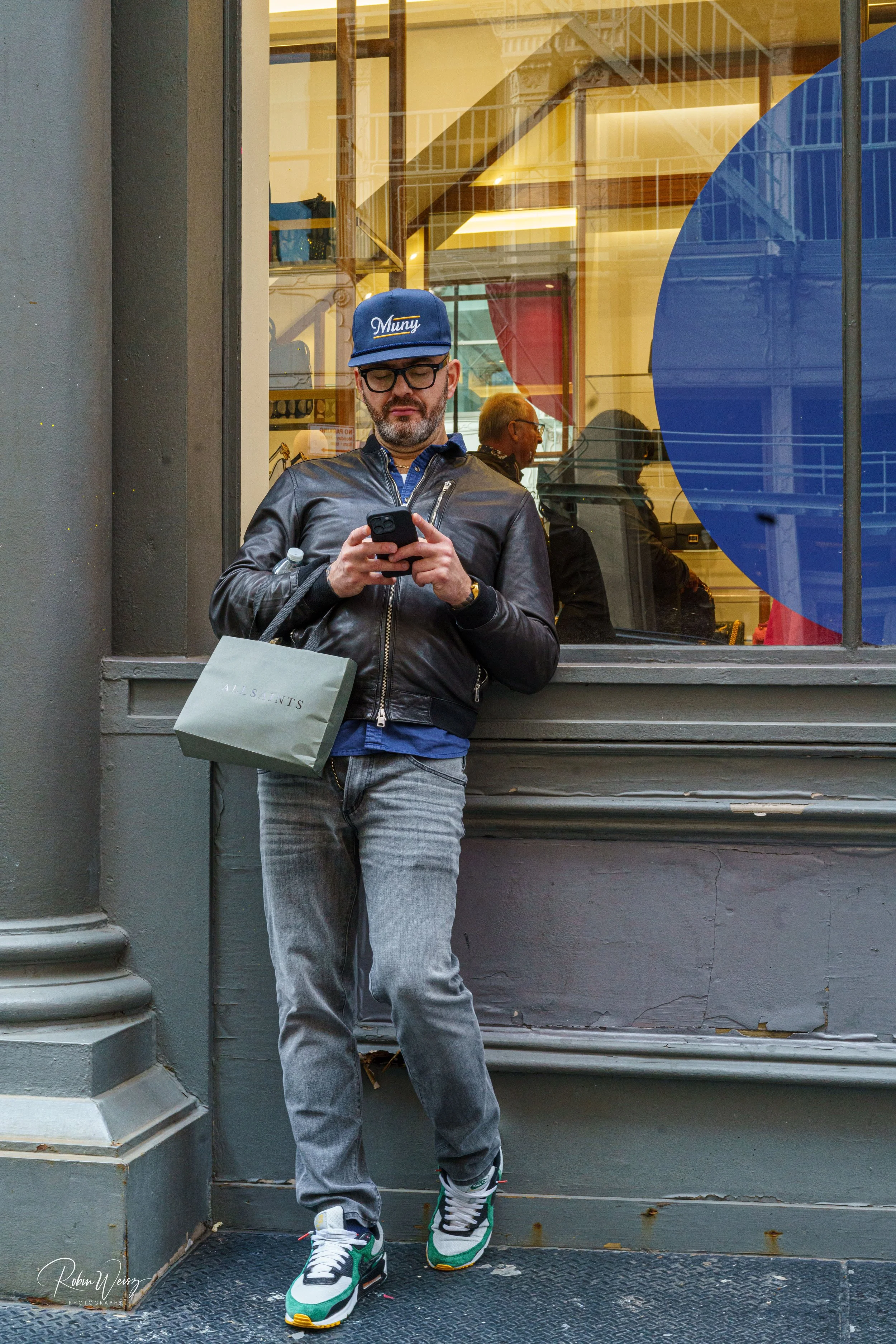 Man in a blue cap and leather jacket using a smartphone outside a storefront, holding an AllSaints shopping bag.