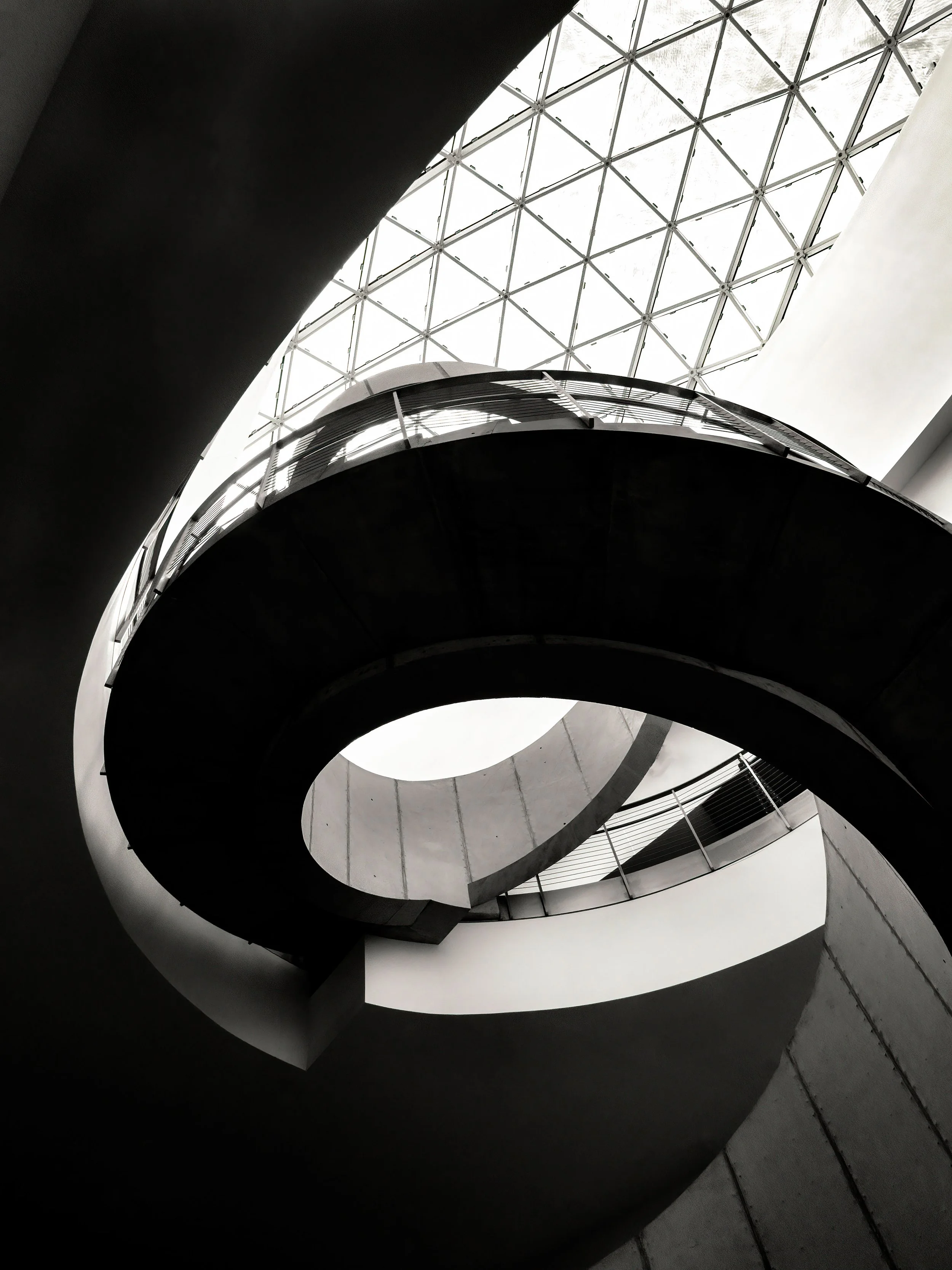 Black-and-white upward view of a modern building interior with curved balconies and a geometric glass dome ceiling