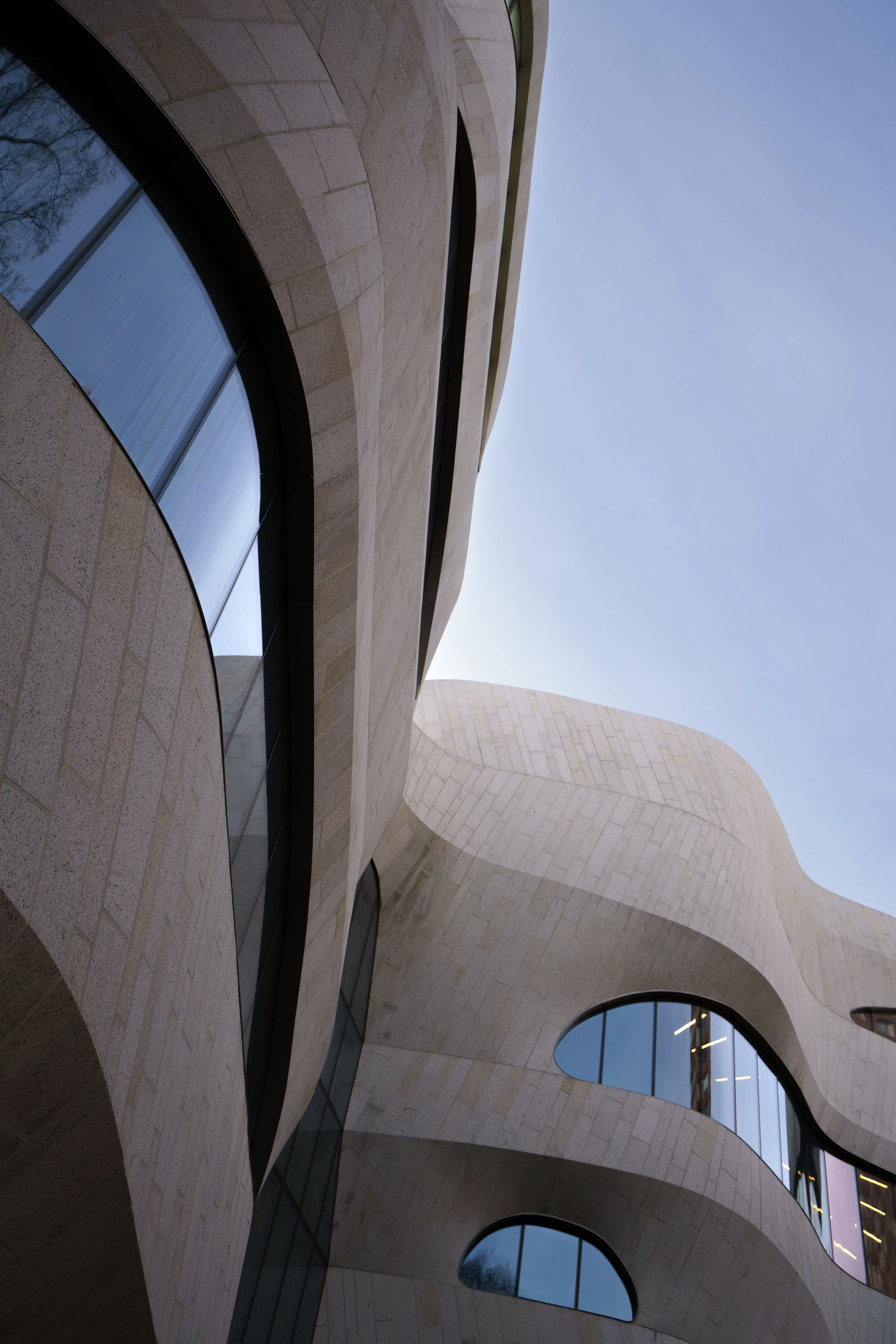 Upward view of a modern stone-clad building with curved arches, large windows, and a blue sky.