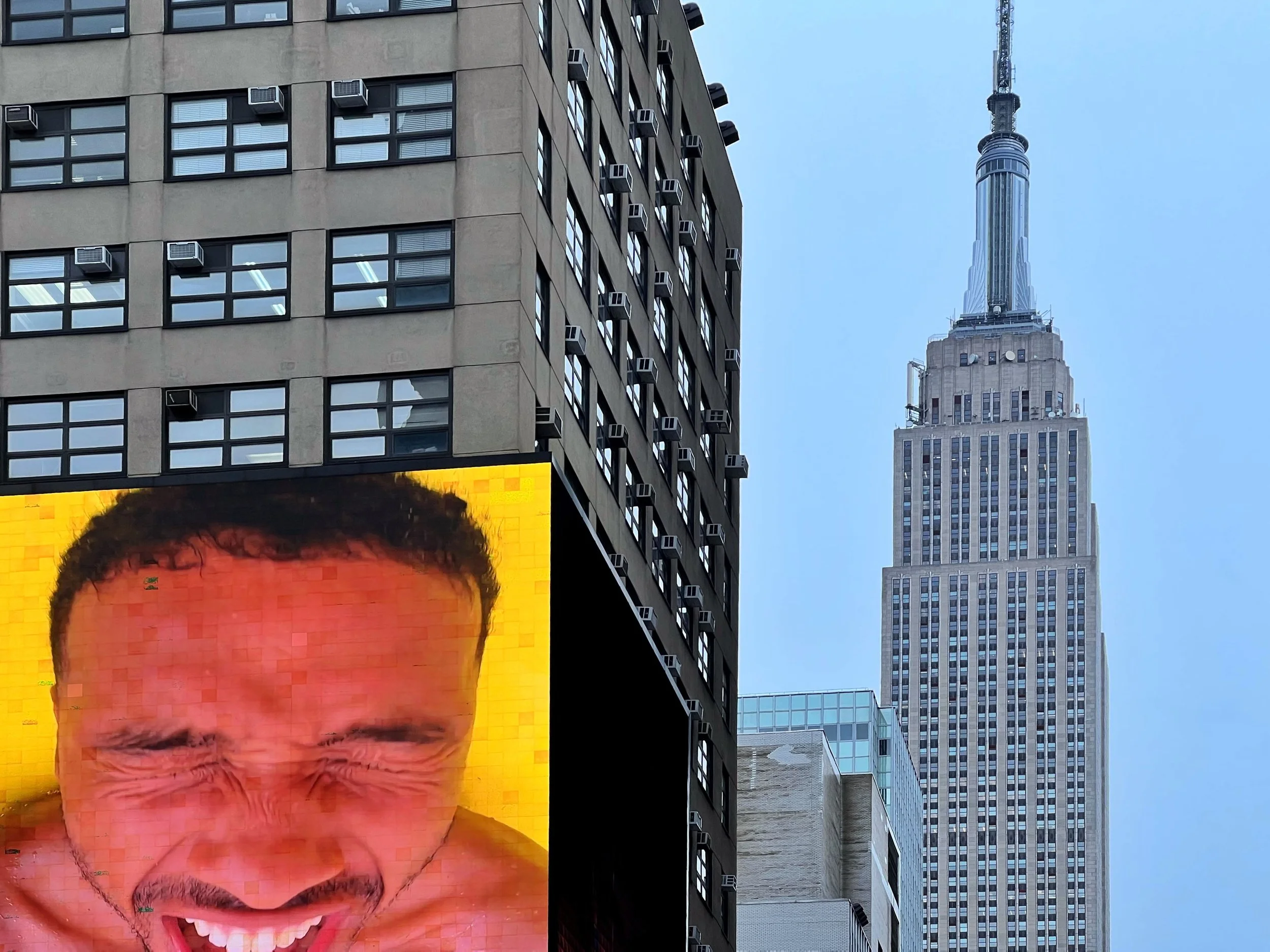 New York City street view featuring a mural portrait, air conditioners on a building, and the Empire State Building under a blue sky