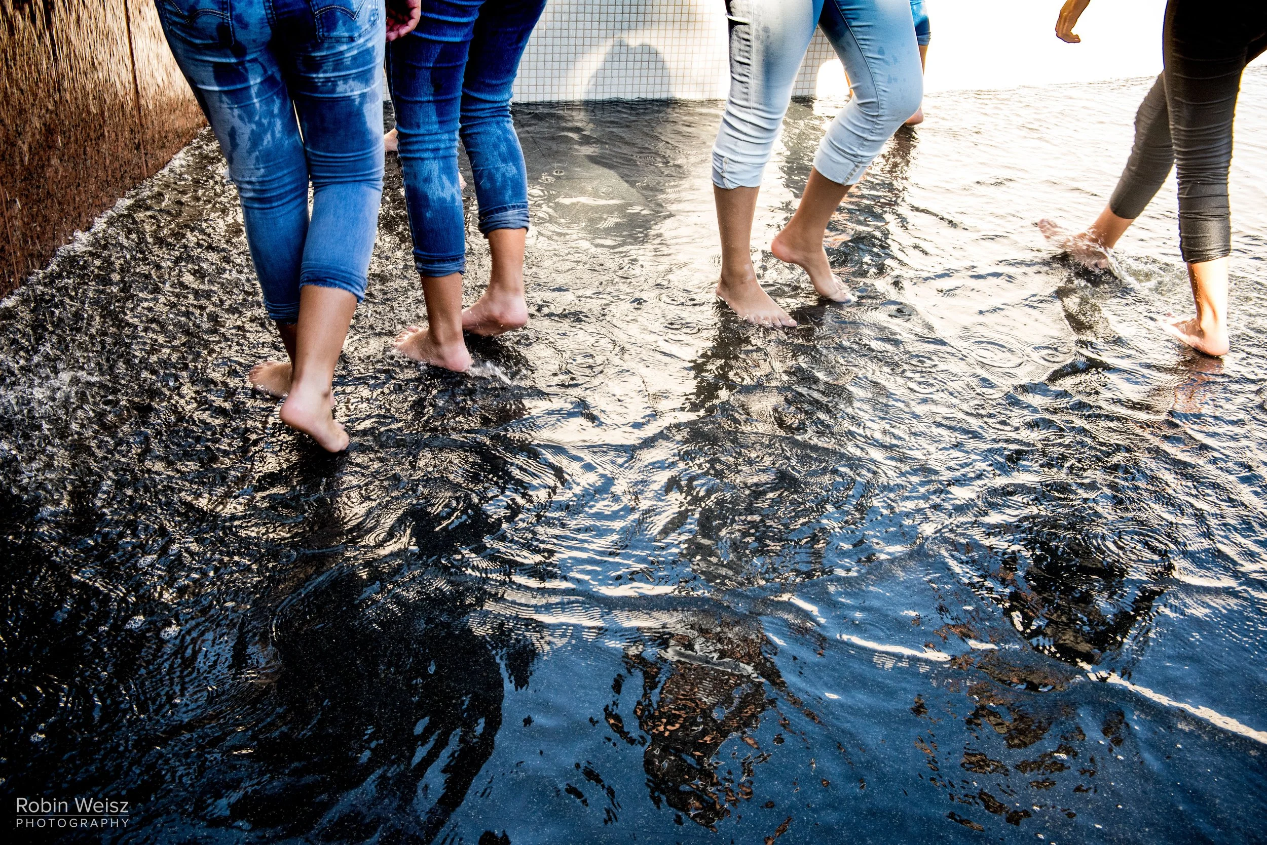 People in rolled-up jeans walking barefoot through shallow water on a rocky shoreline.
