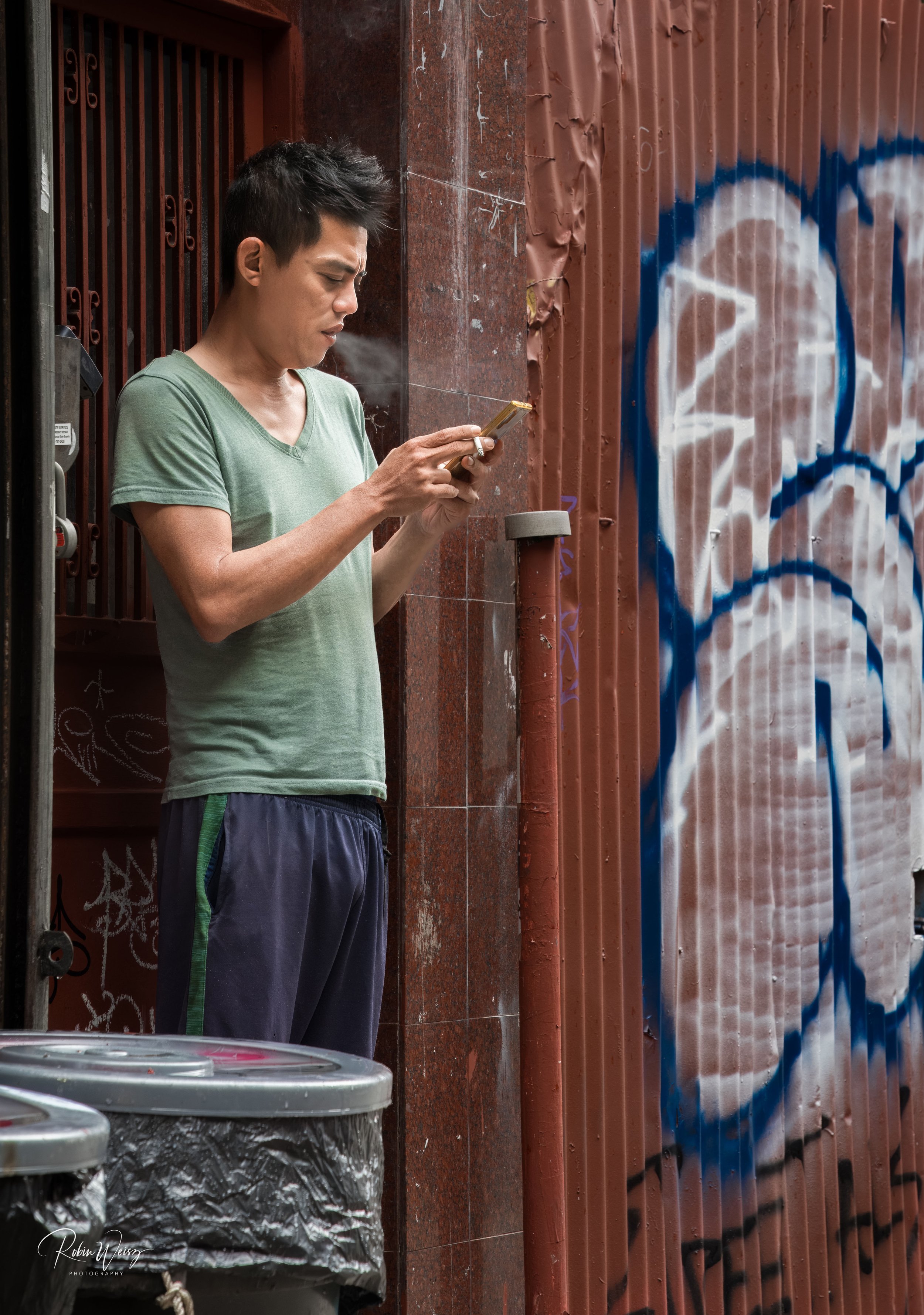 Man in a green shirt using a smartphone while smoking outside a graffiti-covered building