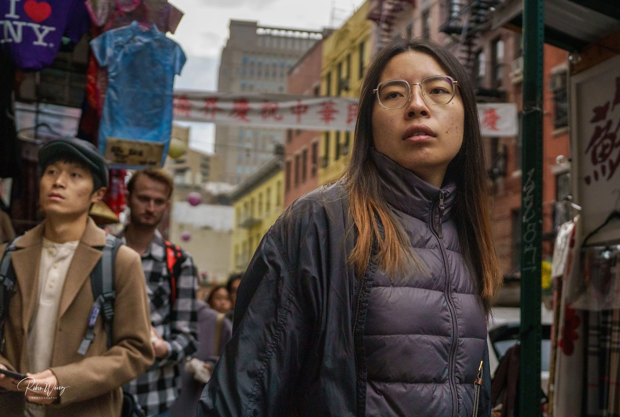Young woman wearing glasses and a black puffer jacket in a busy Chinatown street market, with pedestrians and Chinese signs in the background