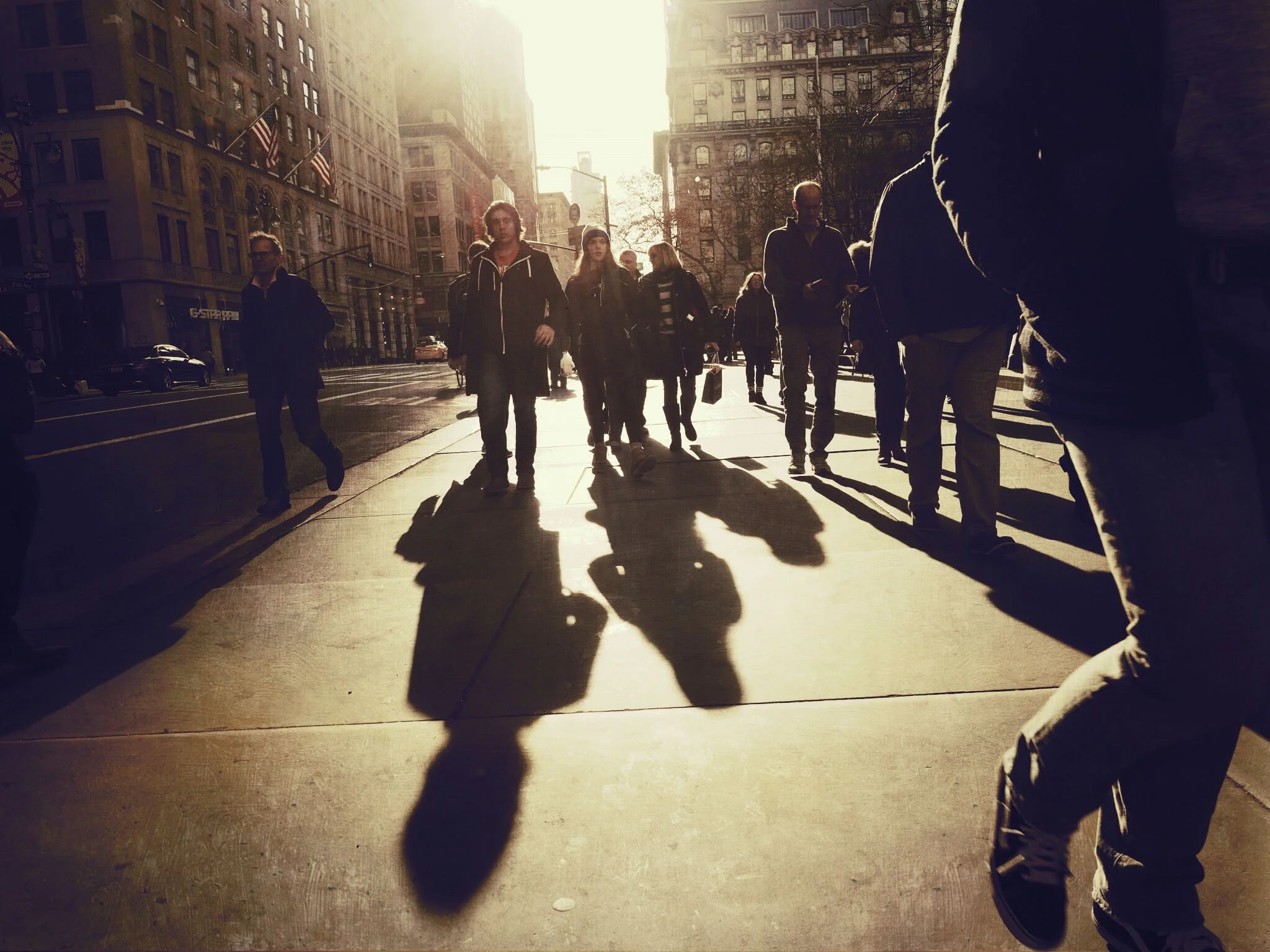 Pedestrians walking along a sunlit downtown city sidewalk beside historic buildings and American flags.