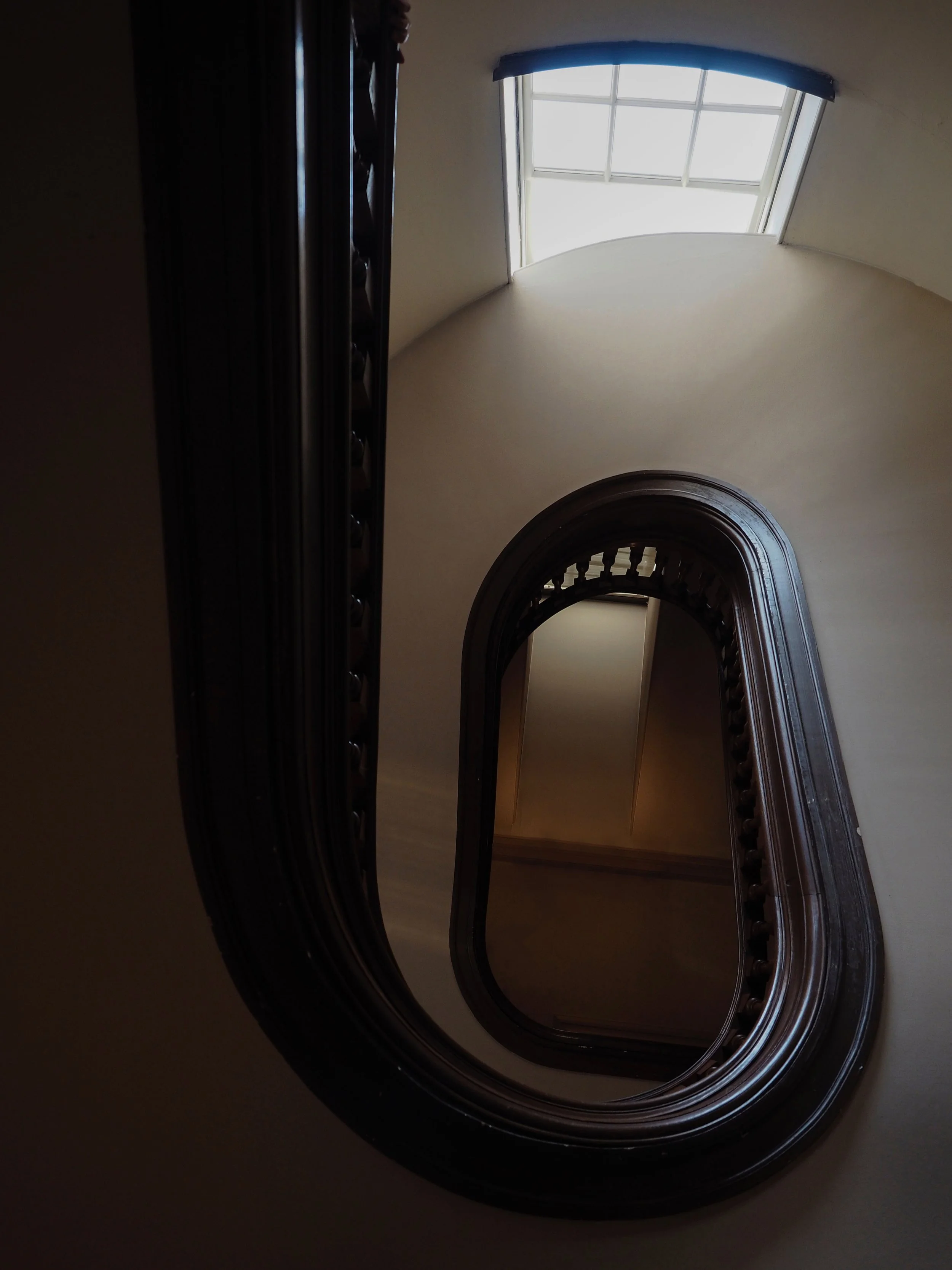 Interior view of a curved wooden staircase with ornate dark railing and a bright multi-pane window