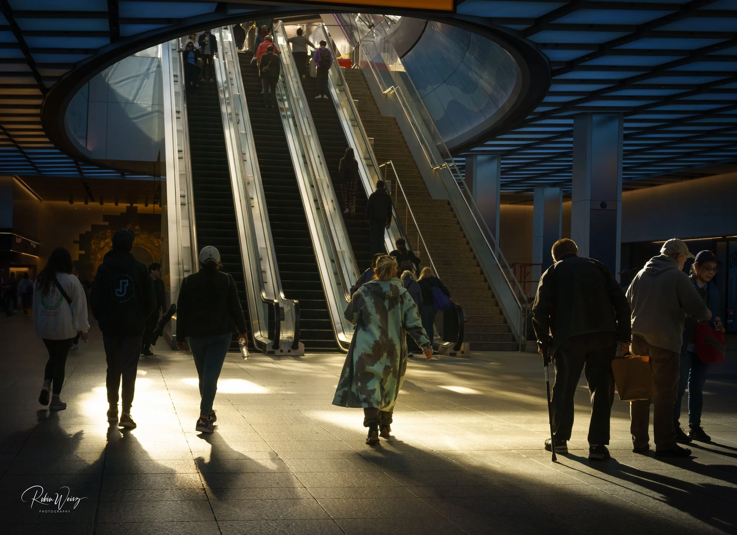 Crowded modern transit station interior with commuters using escalators and stairs beneath a blue-lit geometric ceiling