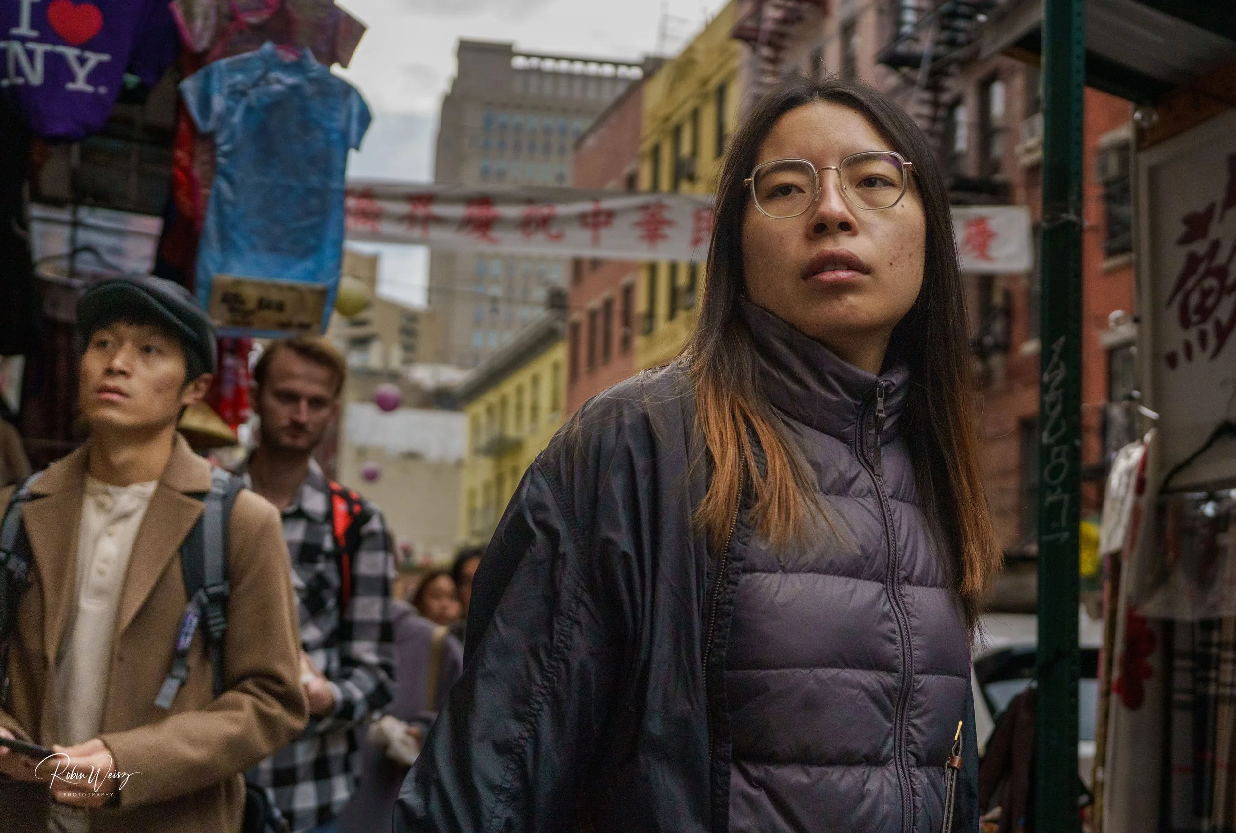 Woman wearing glasses and a black puffer jacket walks through a busy Chinatown street market.
