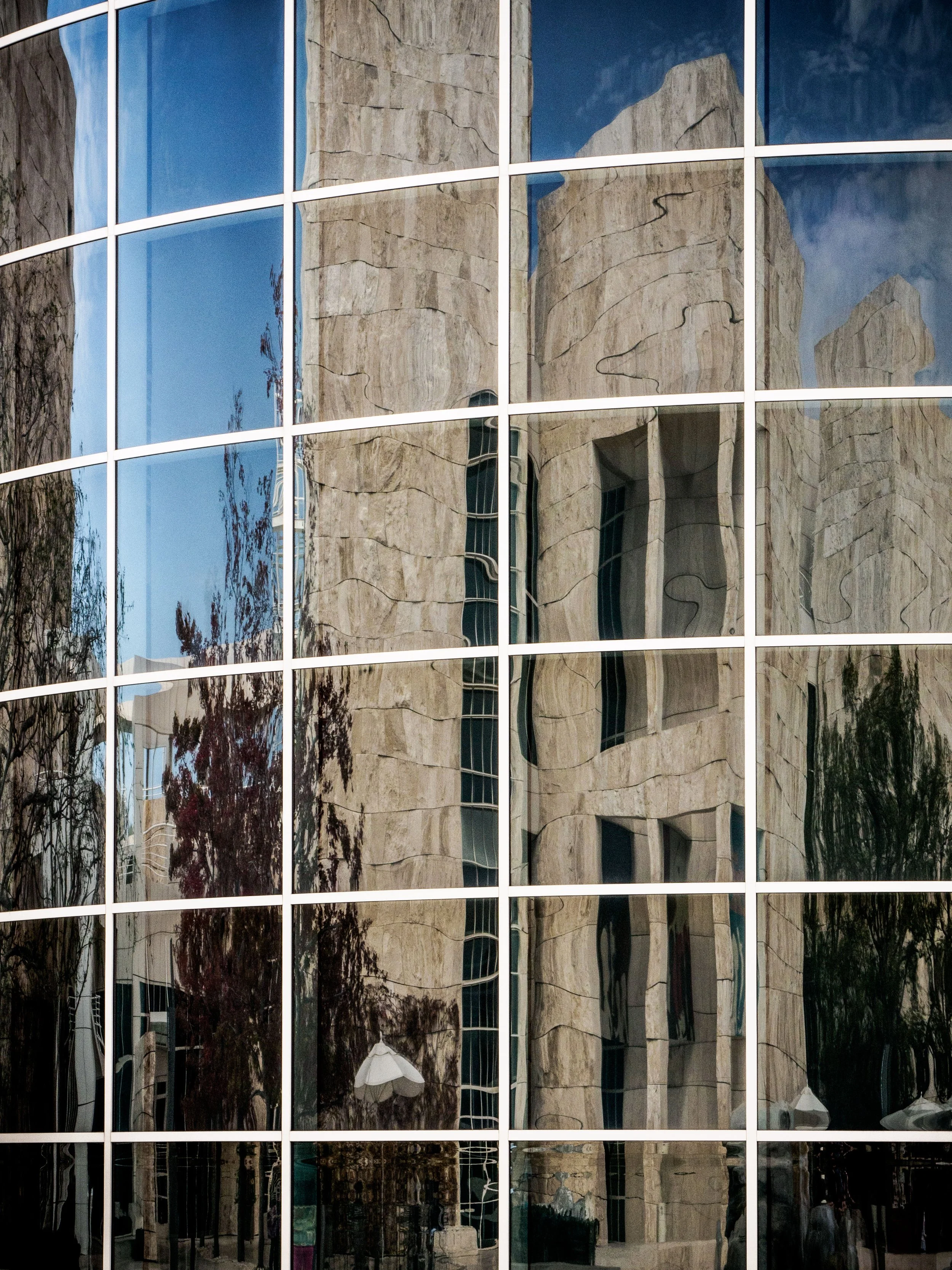 Reflective glass building facade mirroring a stone building, blue sky, and trees