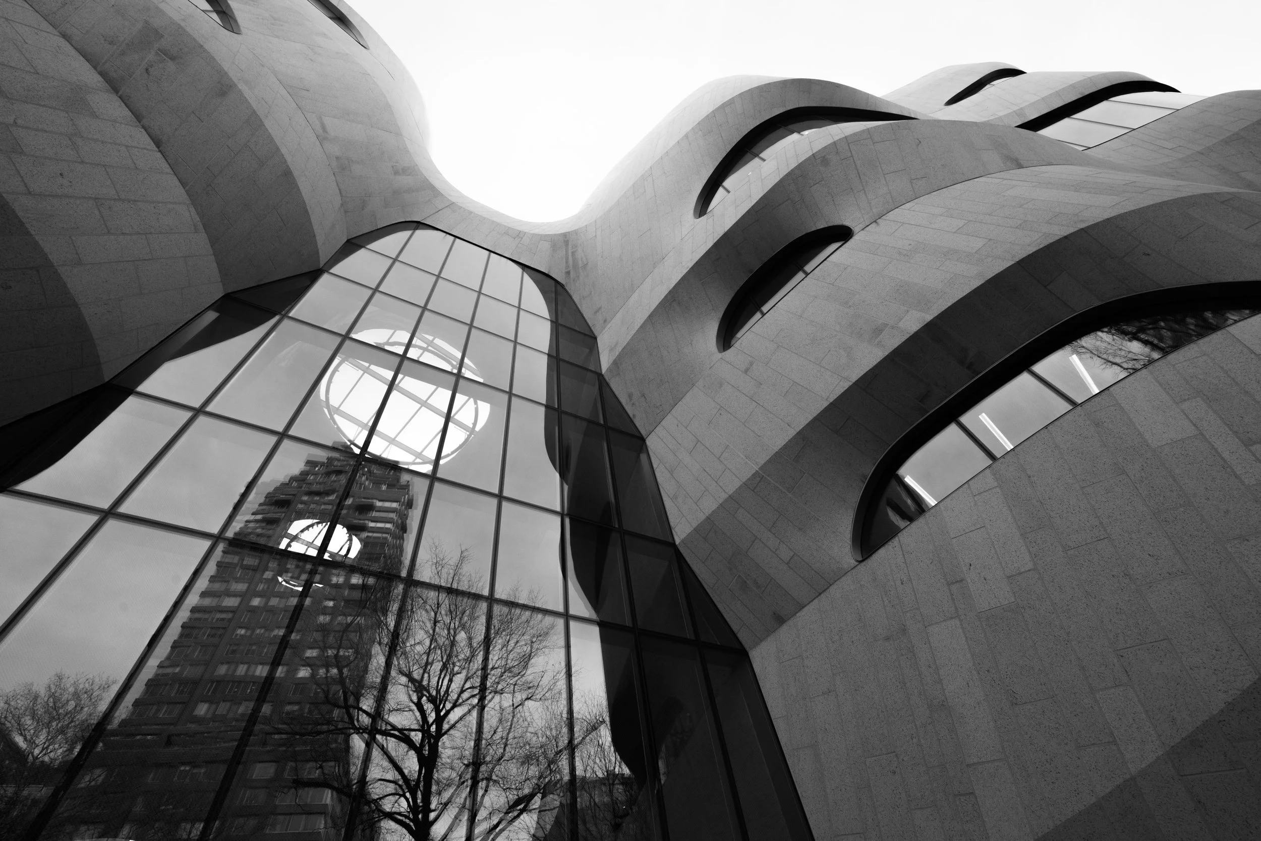 Black-and-white upward view of a modern building with sweeping curved stone walls, glass windows, and reflections of trees and apartments.