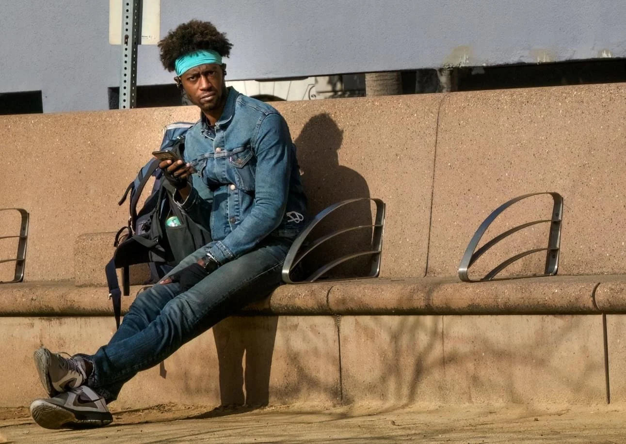 Man wearing a turquoise headband and denim outfit sits on a concrete bench while holding a smartphone and backpack.