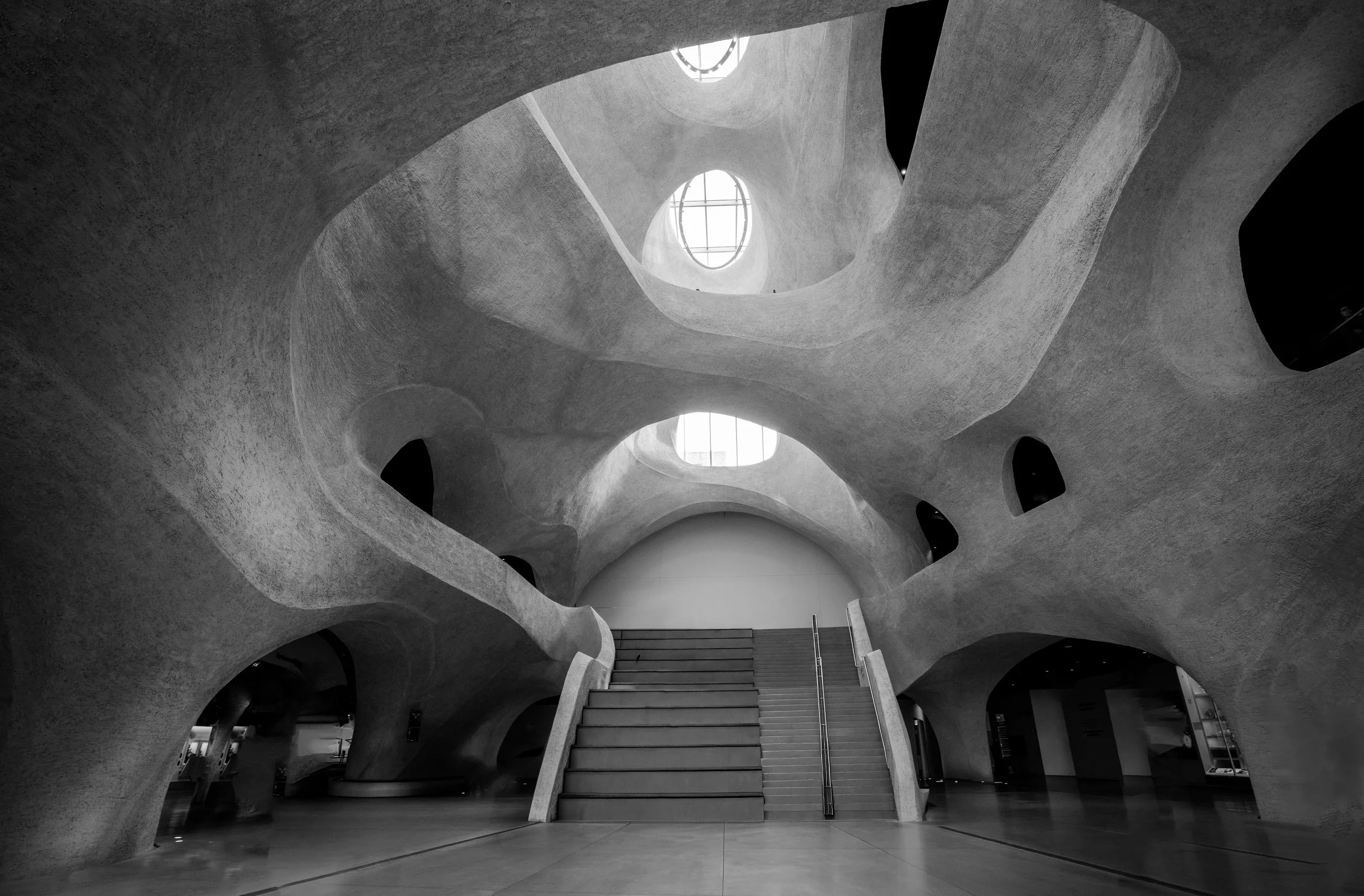 Black-and-white interior of an organic concrete building with sculptural arches, skylights, and a central staircase