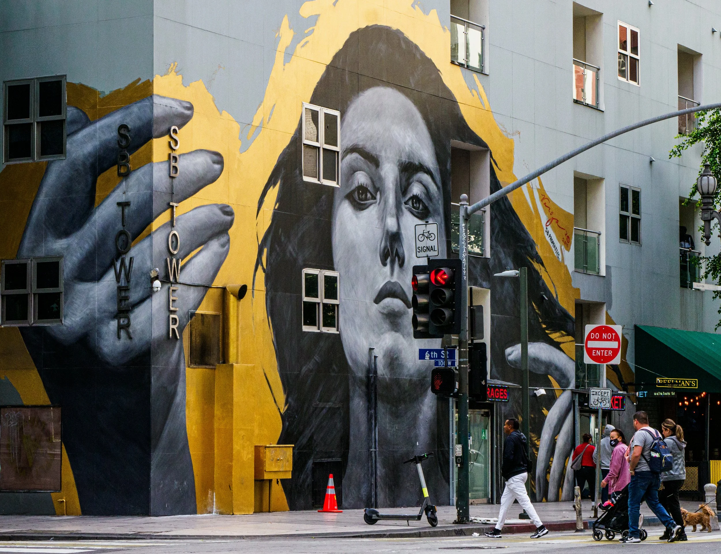 Large black-and-white mural of a woman and raised hands on a downtown building at 6th Street, with traffic lights and pedestrians in the foreground.