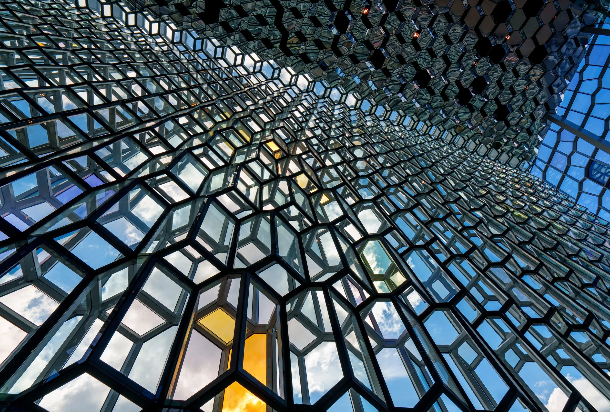 Abstract upward view of a geometric glass ceiling with black honeycomb frames reflecting blue sky and clouds