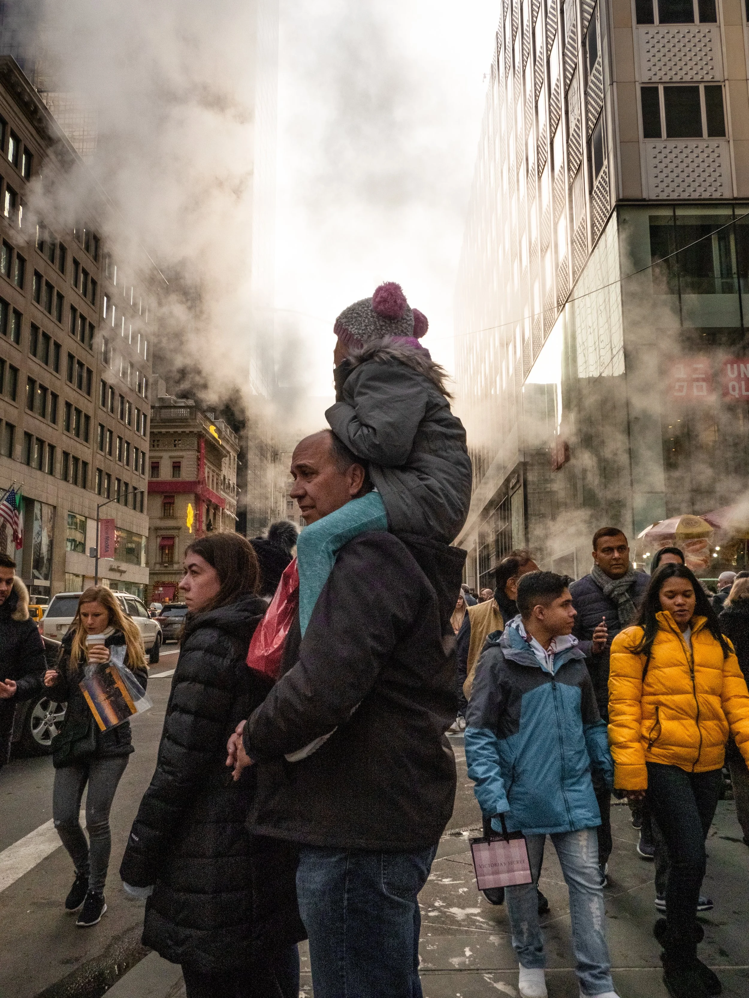 Crowded city street with a parent carrying a child on their shoulders between tall office buildings and steam rising from the sidewalk.