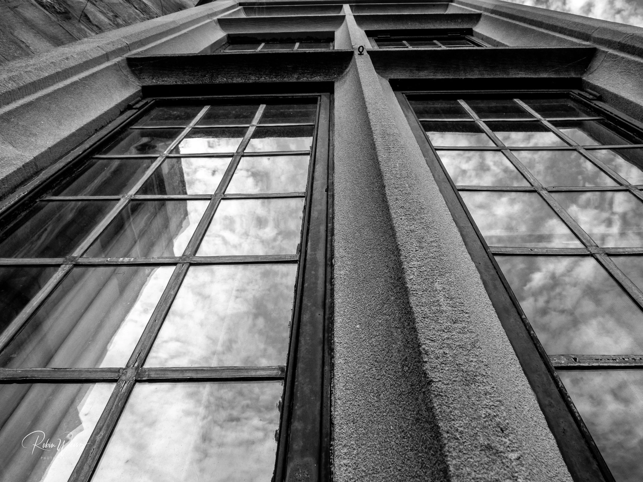 Black-and-white upward view of a historic building facade with tall divided-pane windows reflecting cloudy skies