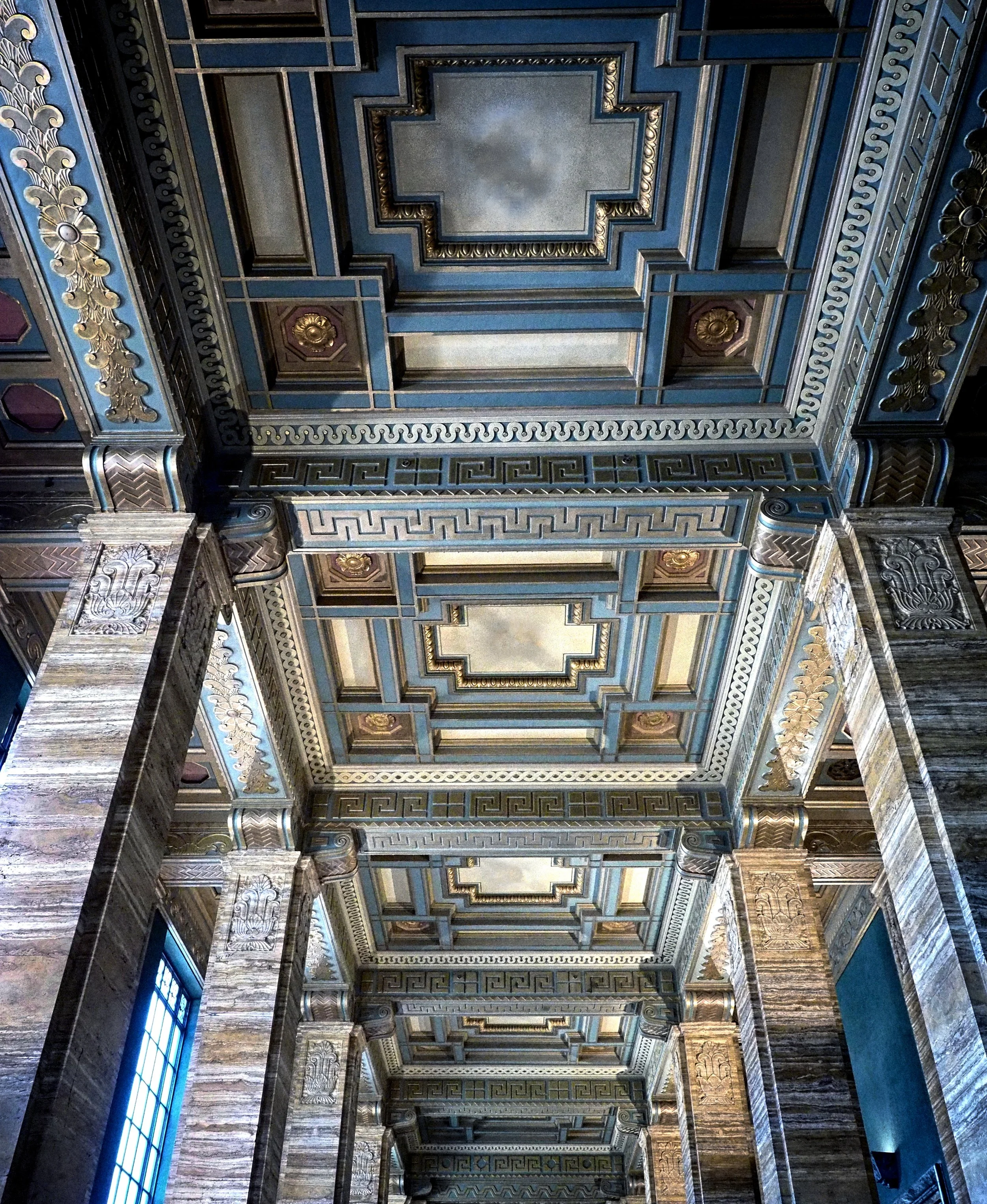 Ornate Art Deco interior ceiling with geometric panels, Greek key patterns, gold floral details, and stone columns