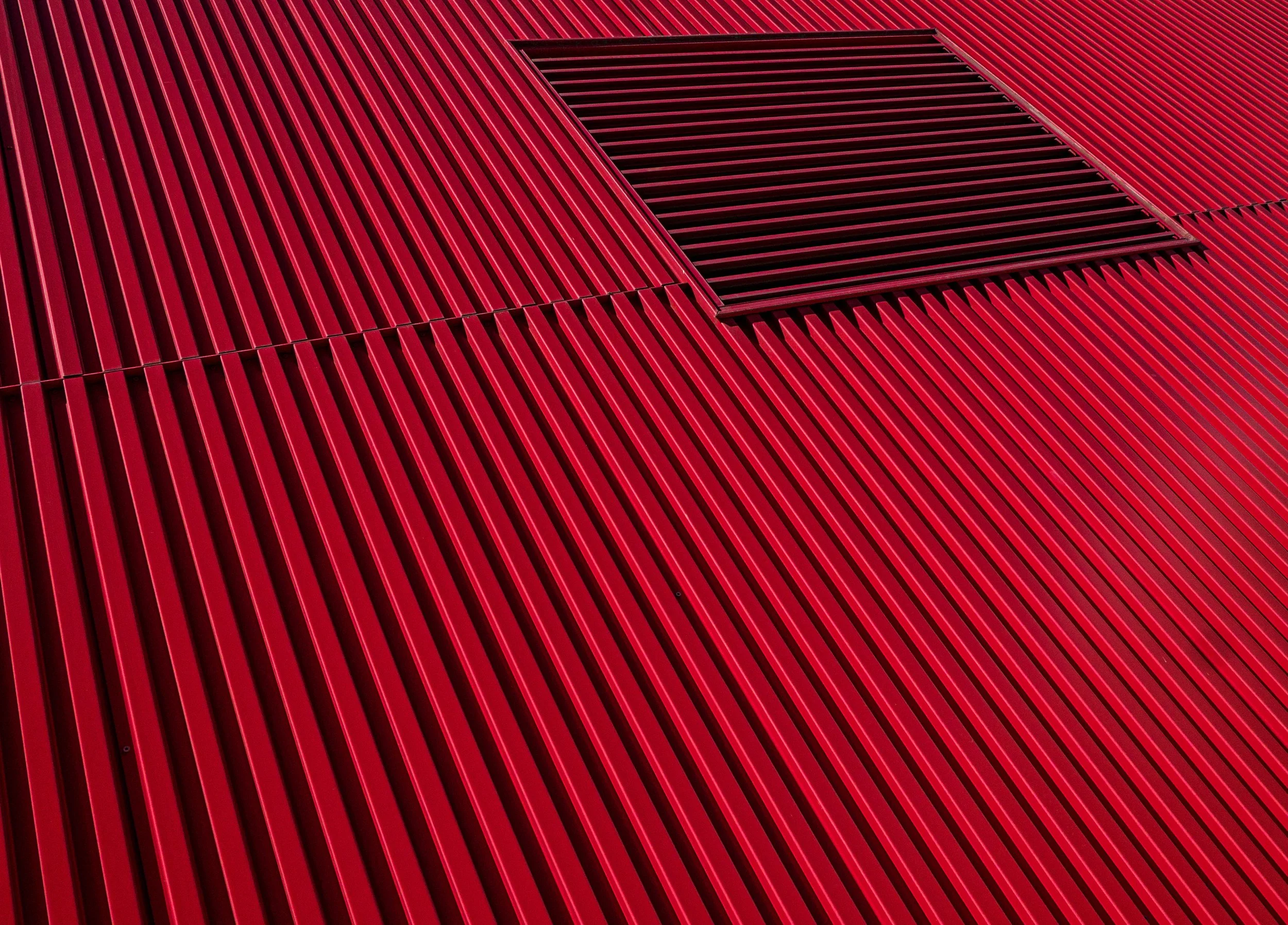 Abstract close-up of red corrugated metal panels with a dark rectangular vent or grille