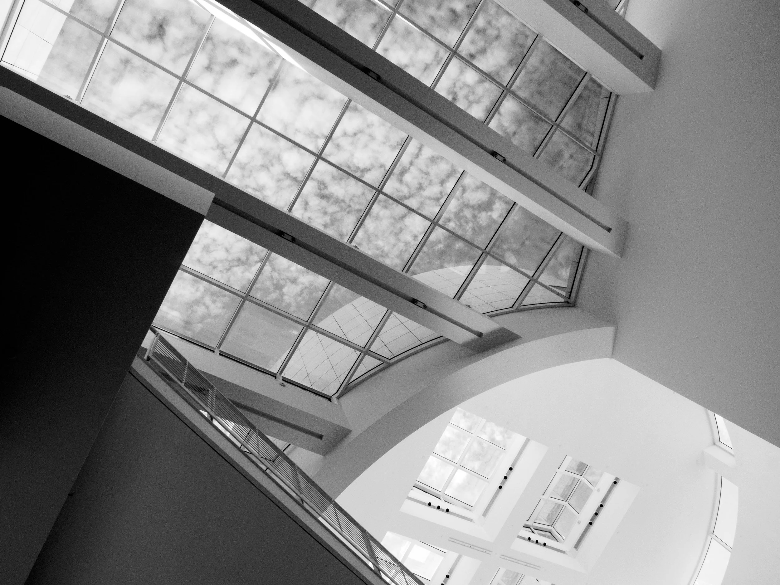 Black-and-white view of a modern atrium with geometric skylights, exposed beams, and balconies beneath a cloudy sky.