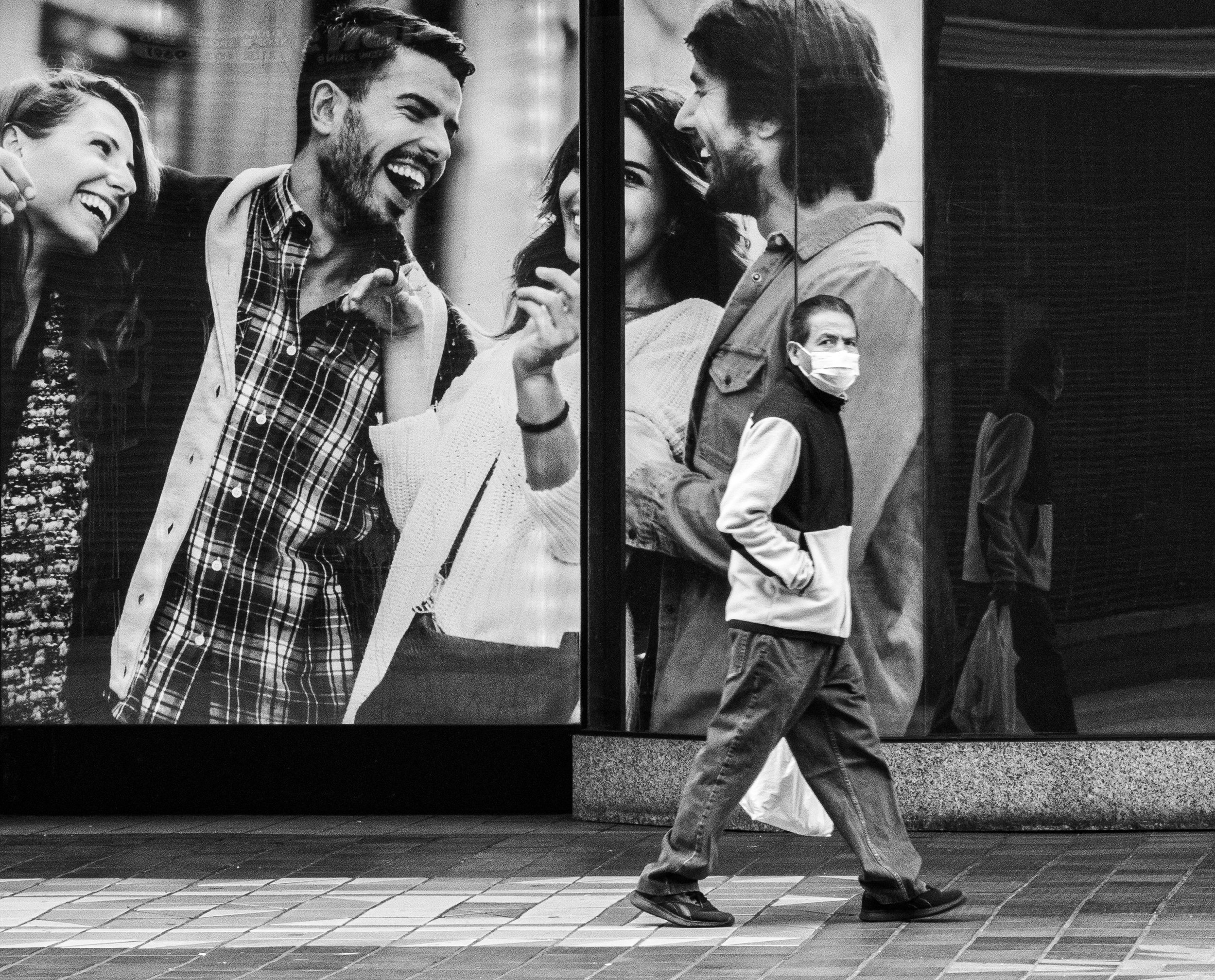 Black-and-white street scene showing a masked man walking past a storefront window with oversized laughing couple portraits.