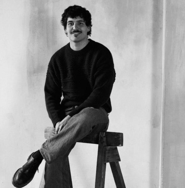 Black and white photo of a man with curly hair and a mustache, sitting on a wooden stool against a plain wall. He is wearing a dark sweater, light jeans, and black shoes, looking at the camera.