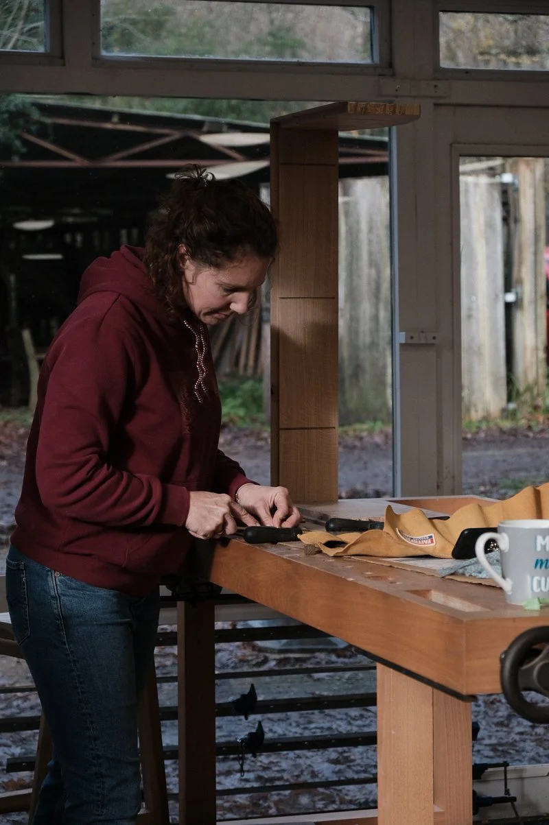 A woman in a maroon hoodie is working on a woodworking project at a workbench, cutting wood with a hand saw in a workshop or garage.