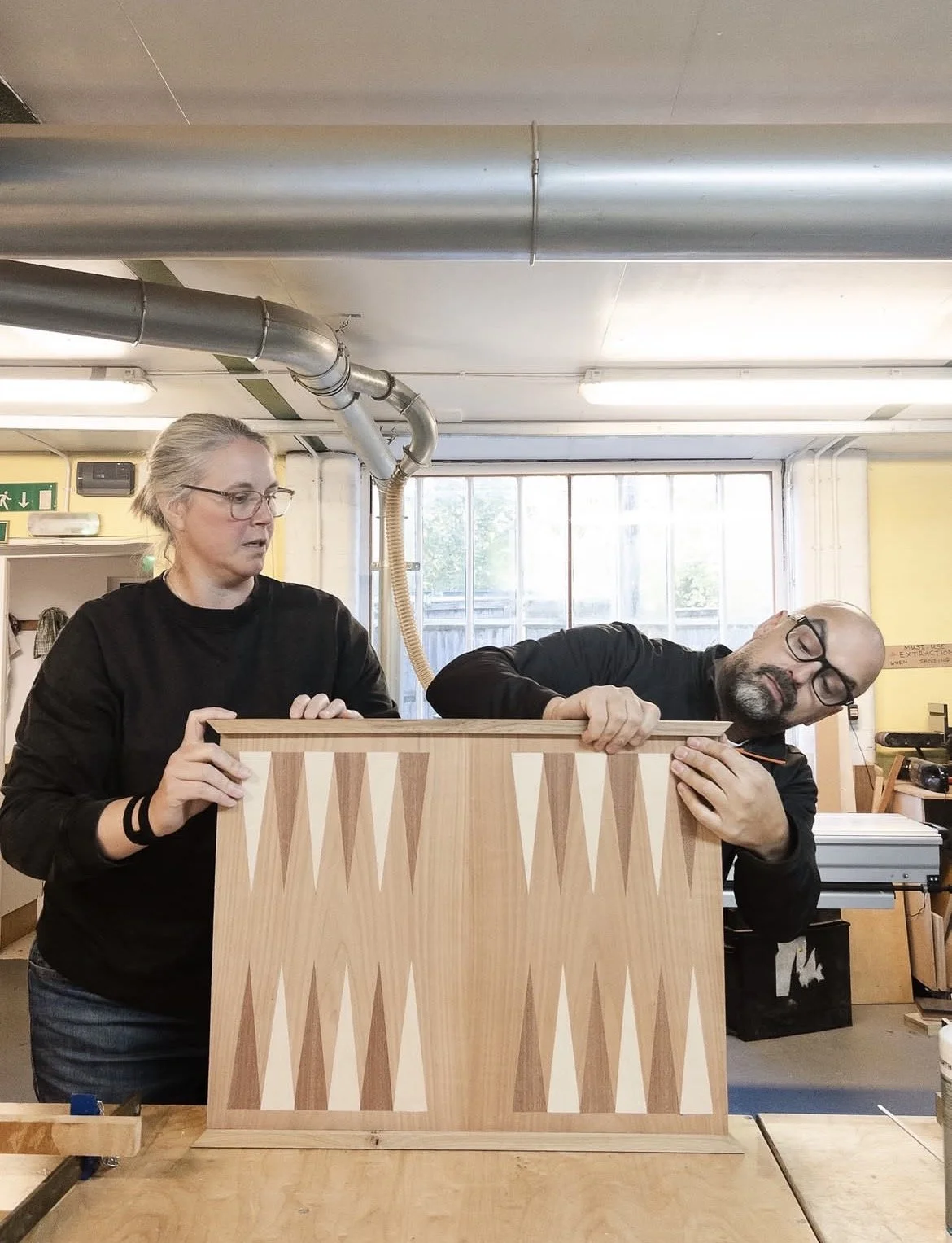 A woman and a man are working on a wooden chessboard in a woodworking workshop. The woman is holding the chessboard upright, looking at it, while the man, leaning on the chessboard, appears to be focused or resting with his eyes closed.