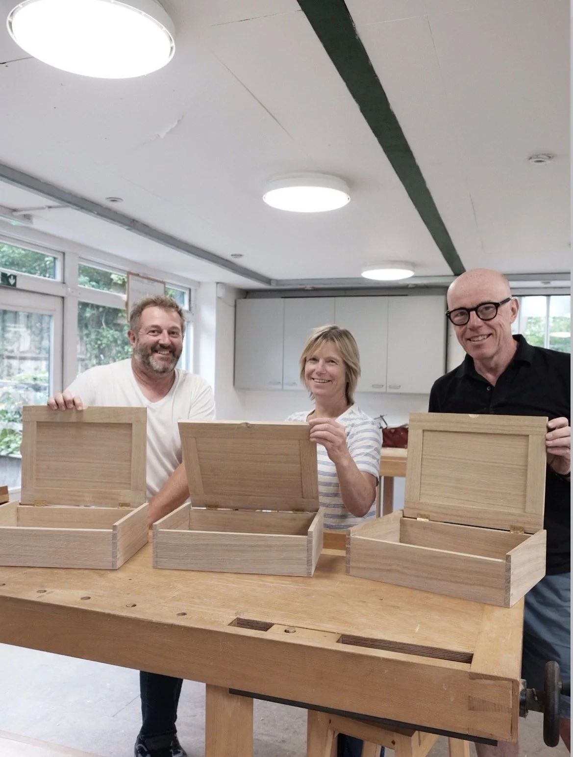 Three people smiling behind open wooden boxes on a workshop table in a room with natural light and overhead ceiling lights.