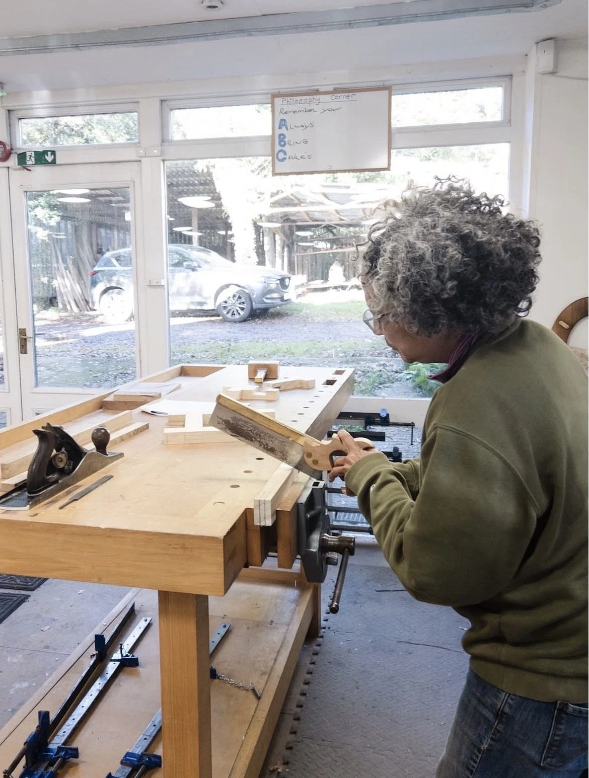 Person with curly gray hair working with a saw in a woodworking shop, with a large window showing cars outside.