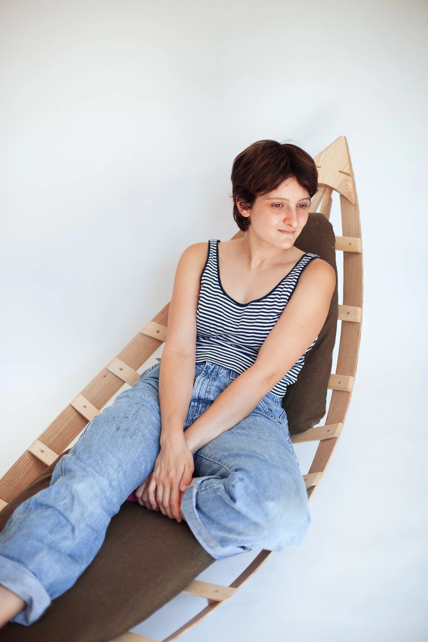 A woman with short brown hair, wearing a black-and-white striped tank top and light blue jeans, sitting on a modern wooden lounge chair with a dark cushion and back support, against a plain white wall.