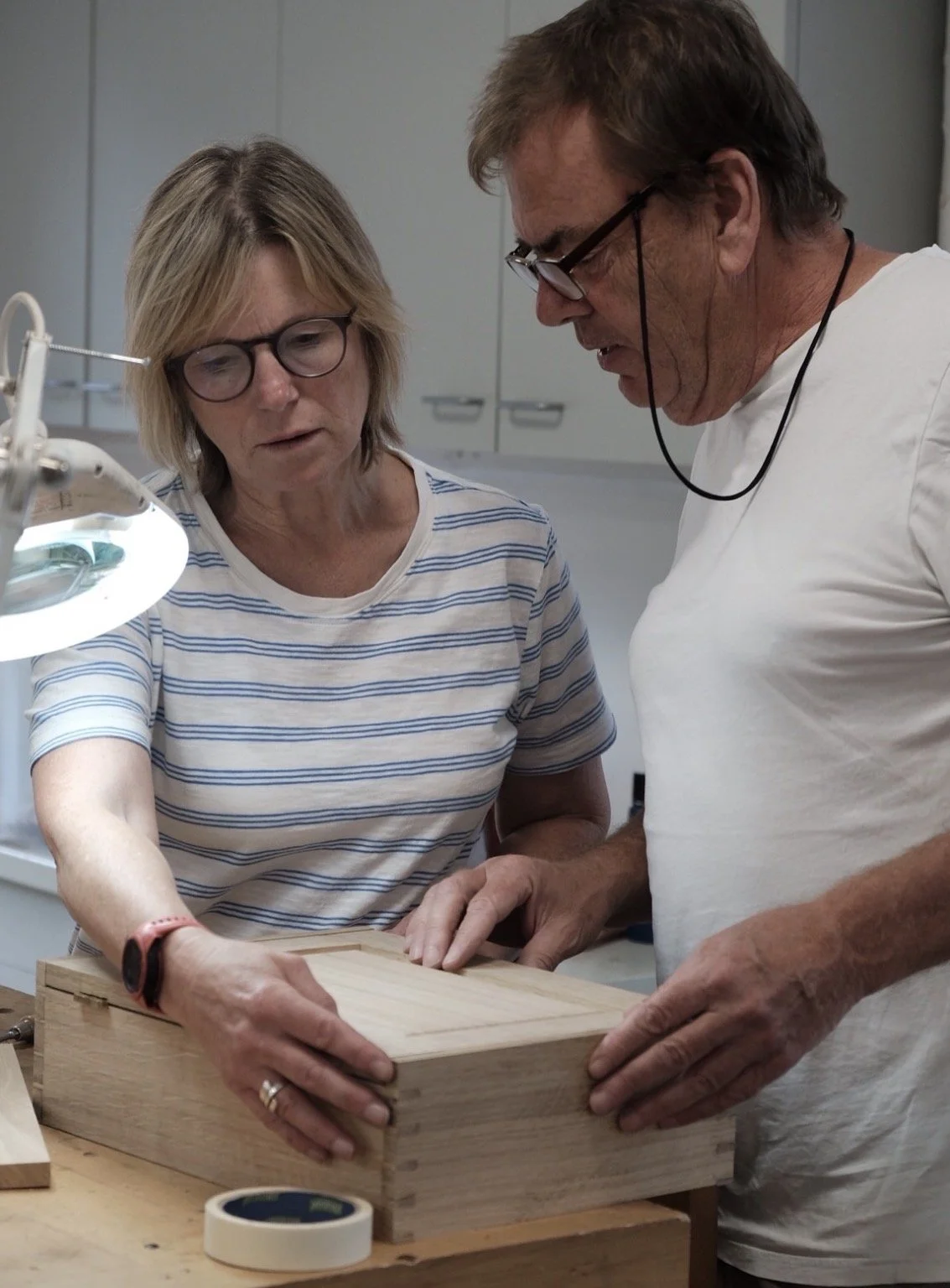 Two people, a woman and a man, examining a wooden box in a workshop or woodworking space. The woman is pointing at it while the man is holding it.