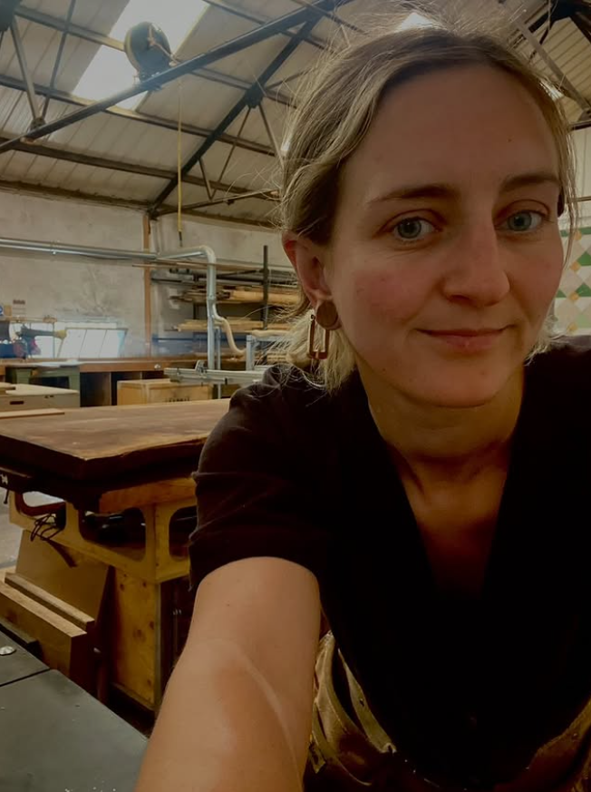 A woman with blonde hair taking a selfie in a woodworking workshop with various tools, workbenches, and equipment in the background.