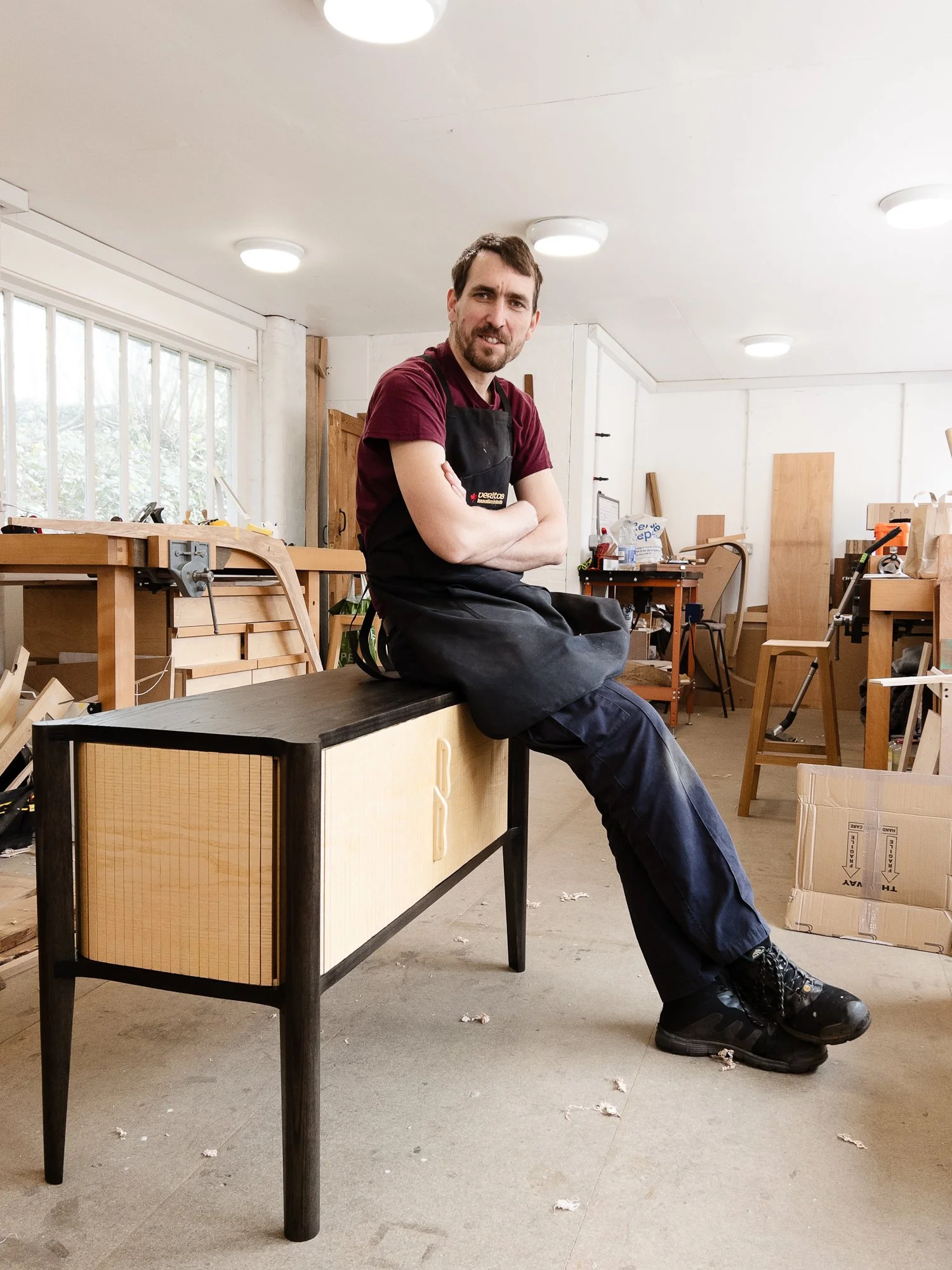 A man sits on a black table in a woodworking shop, wearing a black apron and a maroon shirt. The background has workbenches, tools, and wood pieces, with a large window on the left and a white ceiling with multiple lights.
