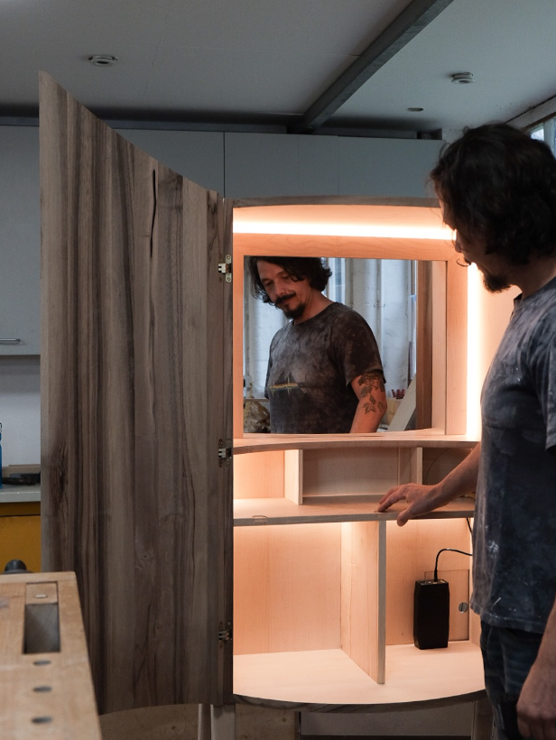A man with dark hair and a beard, wearing a dark T-shirt, is looking at himself in a mirror inside a wooden frame. He is standing in what appears to be a workshop or carpentry area, with unfinished wooden furniture around.