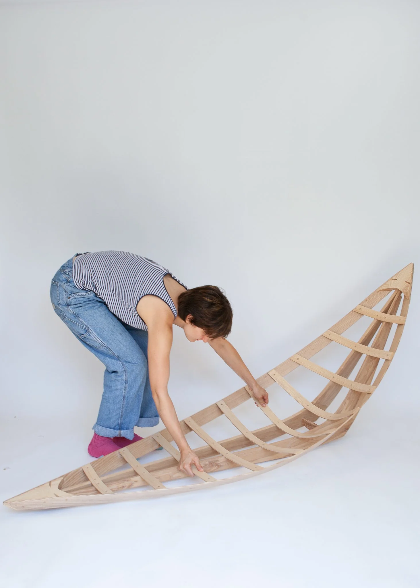 Woman in striped tank top, jeans, and pink socks working on assembling a wooden boat model against a plain white background.