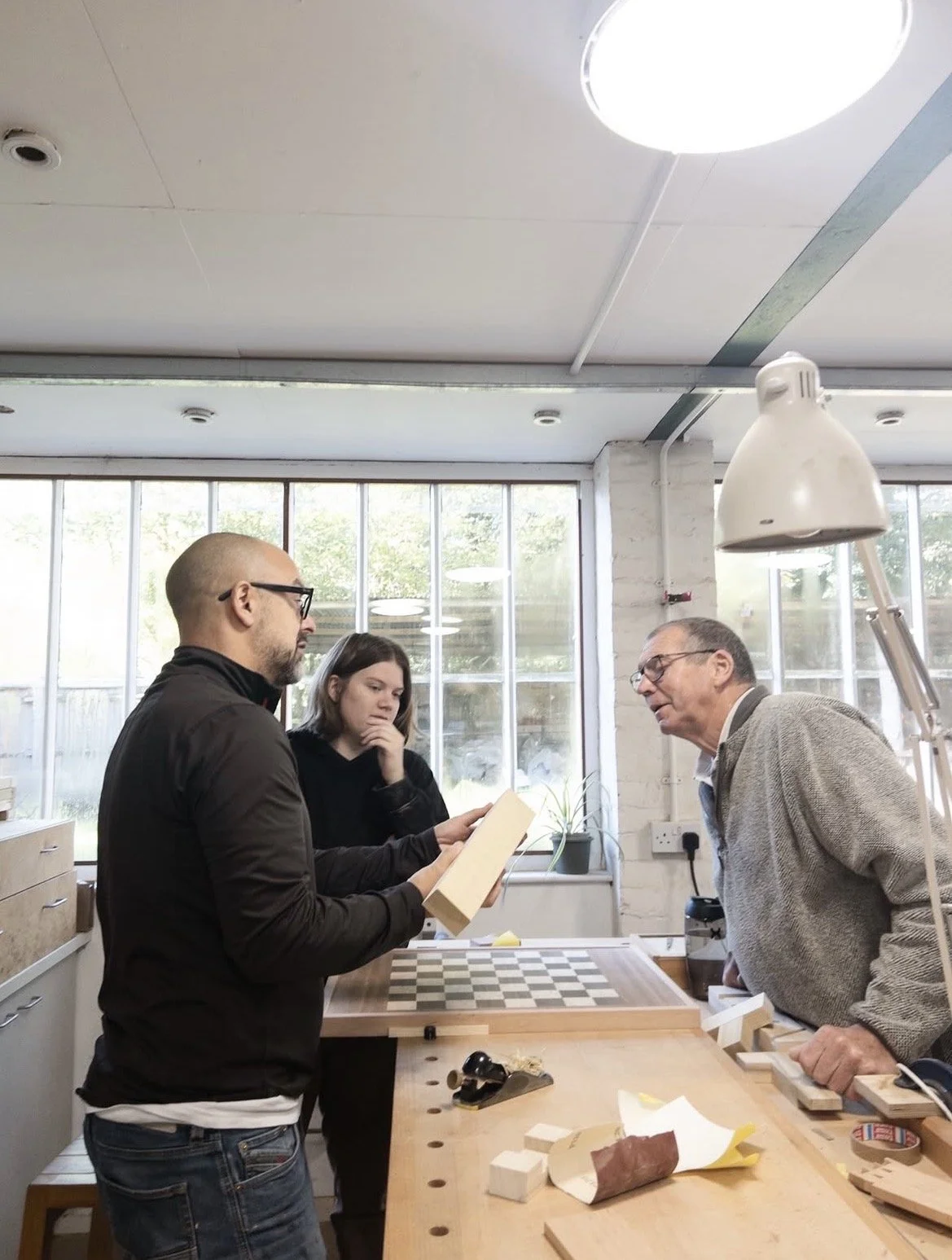 Three people in a brightly lit workshop with large windows, discussing a book and a checkered board. A man with glasses and a woman with a black sweater look on as a younger man in a dark jacket holds the book. An older man in a beige sweater leans on a workbench with various tools and materials.