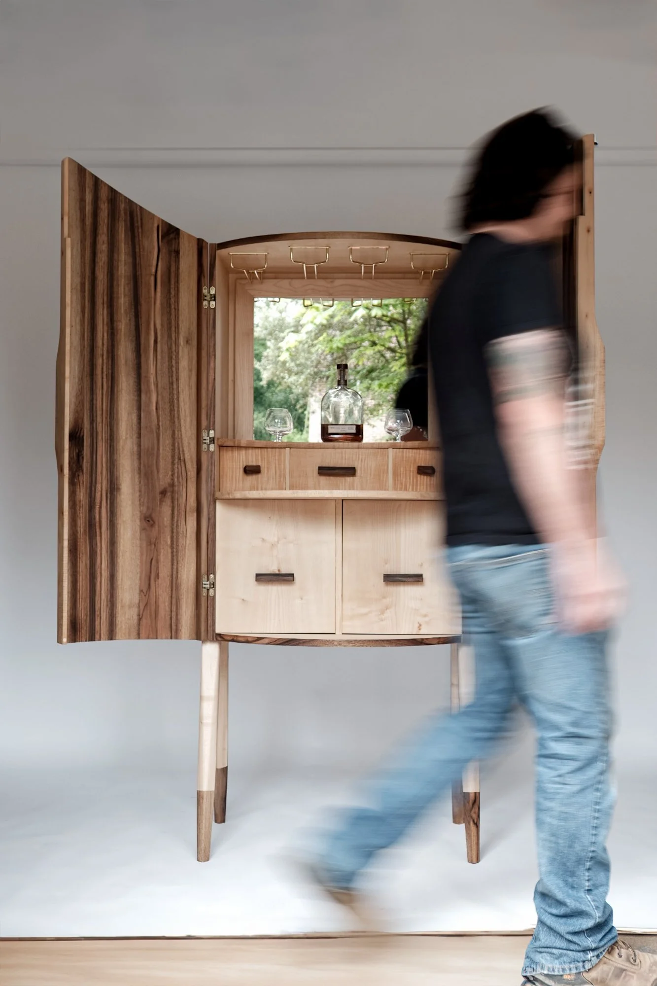 A person walking past a wooden cabinet with open gate, a bottle, and glasses inside, against a light gray wall with a plain flooring.