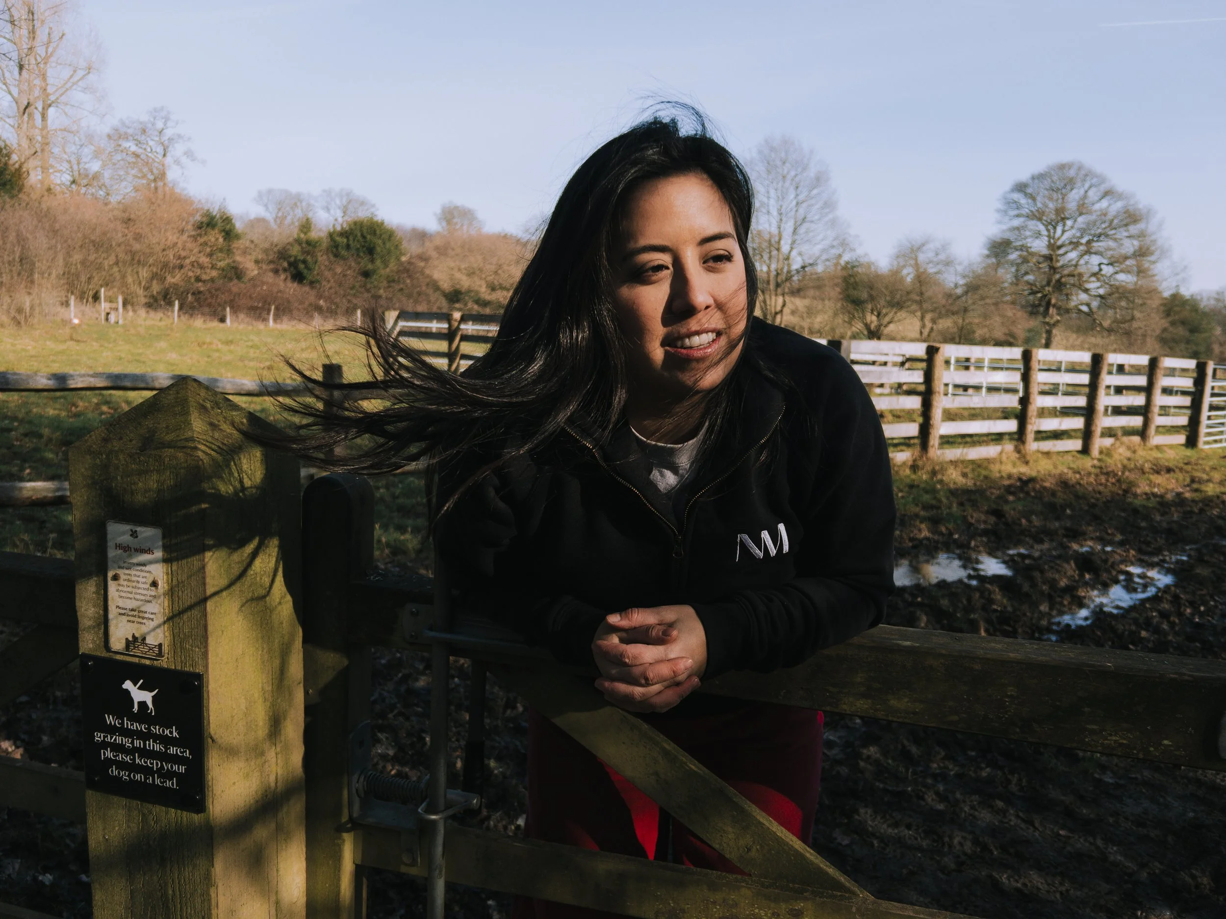 crafts woman leans over public footpath fence at National trust site Scotney castle, her long brown hair floats in the wind as she looks in the middle distance with a half smile.